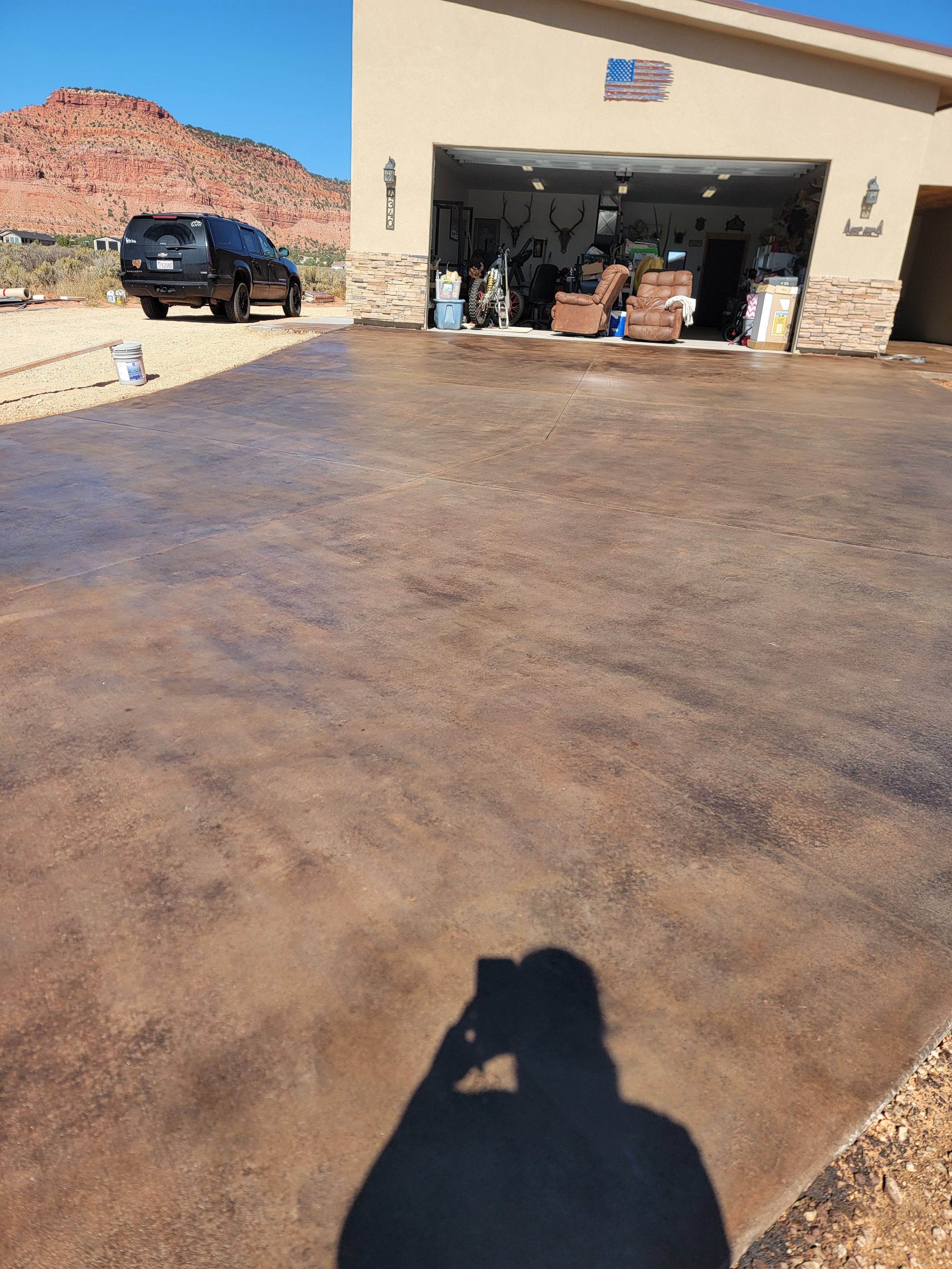 A driveway with both a concrete and gravel section, leading up to a garage on a beige house. The garage is open, showing furniture, deer head mounts, and various household items inside. A black SUV is parked outside, and in the background, there are 