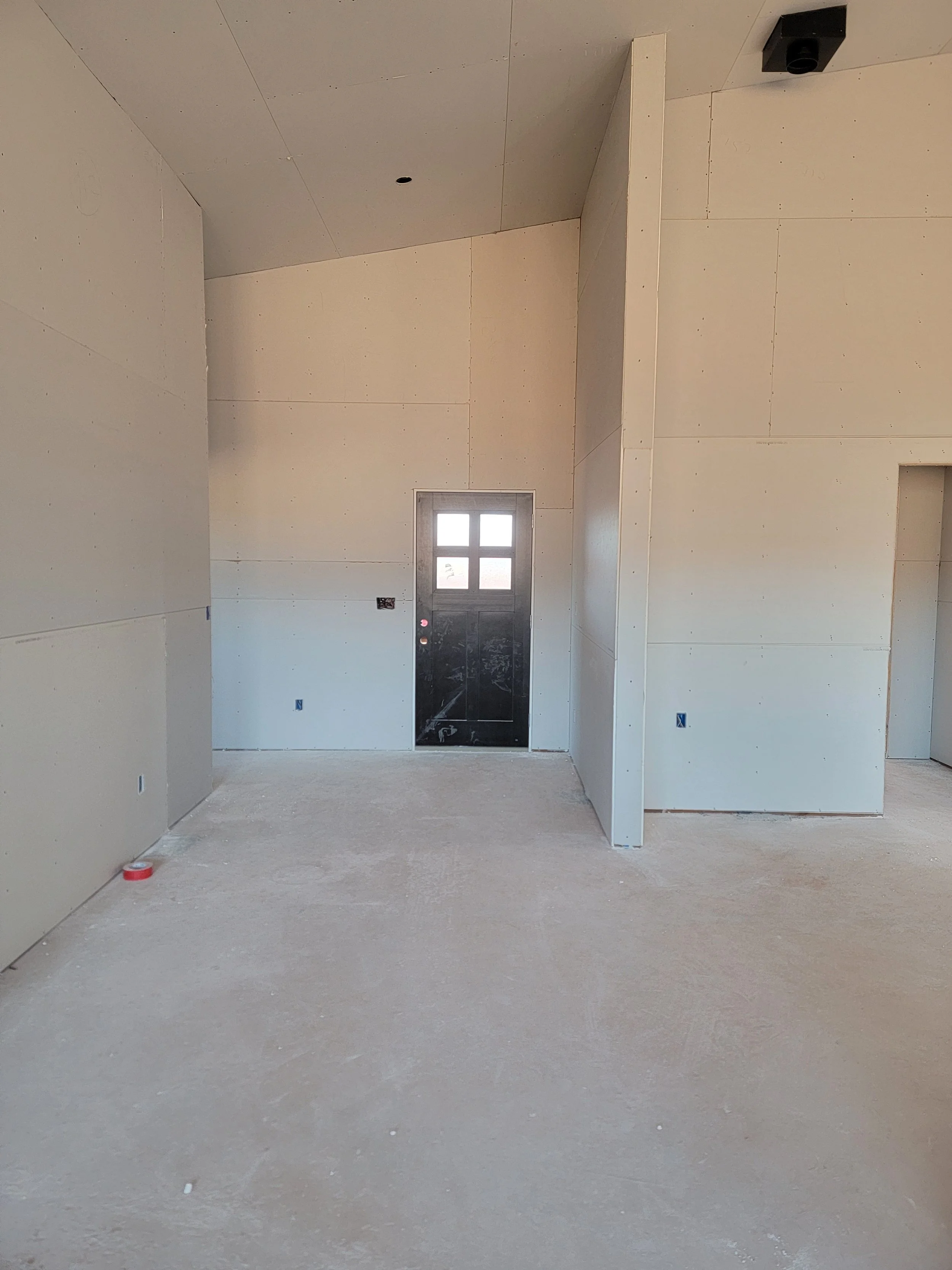 Interior view of a room under construction with unfinished drywall walls, a window at the back, and concrete floor, with a red tape roll on the floor.