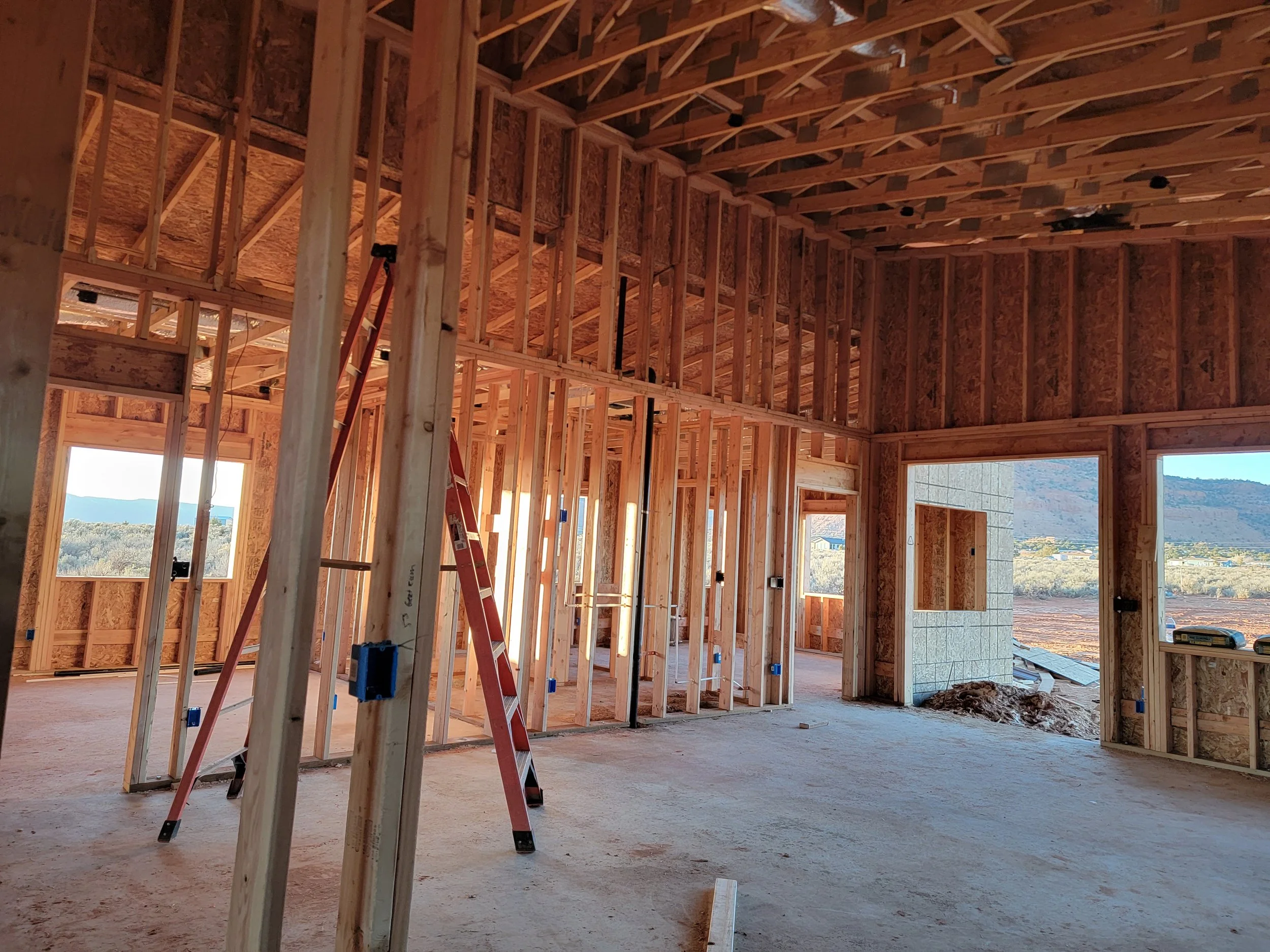 Interior of a house under construction with exposed wooden framing, a ladder, and large windows showing an outdoor view.