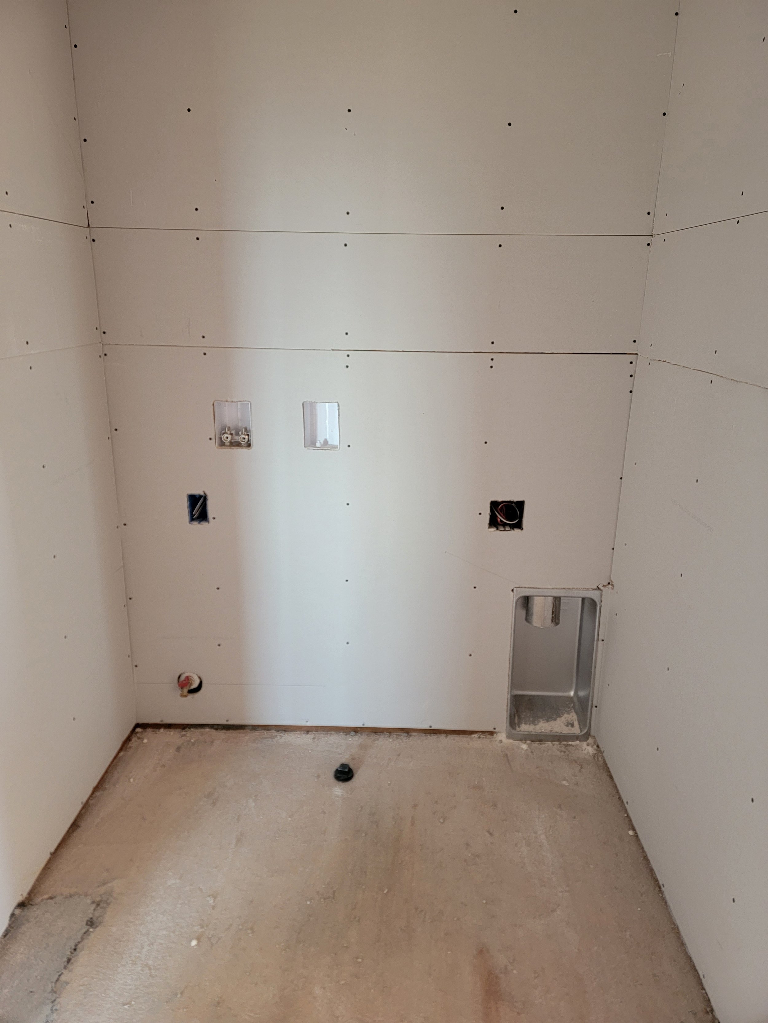 An unfinished room with white drywall walls and a concrete floor, showing electrical wiring boxes and a ventilation opening.