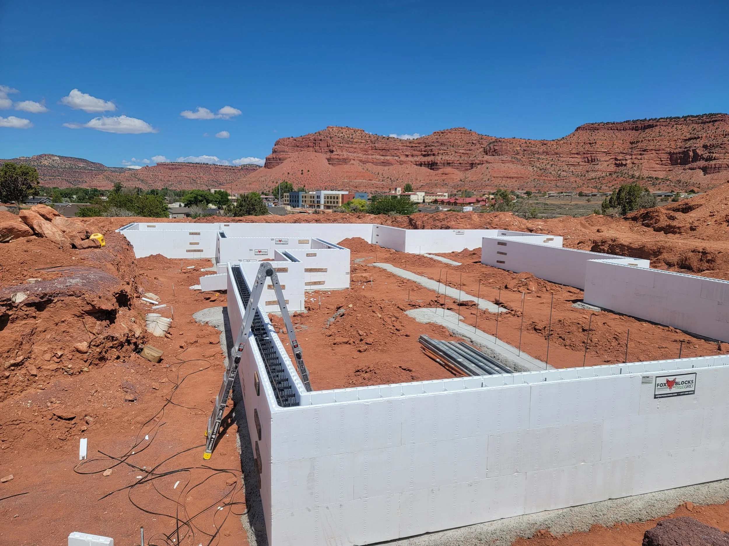 Construction site with white wall panels and red dirt, desert landscape with red rock formations and a blue sky in the background.