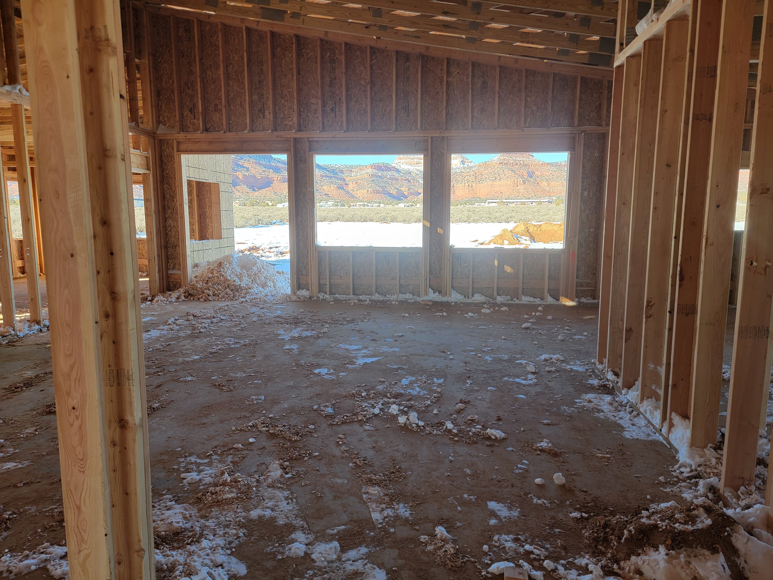 Interior of a house under construction with exposed wooden framing, large windows showing a mountainous landscape, wood debris on the floor, and snow outside.