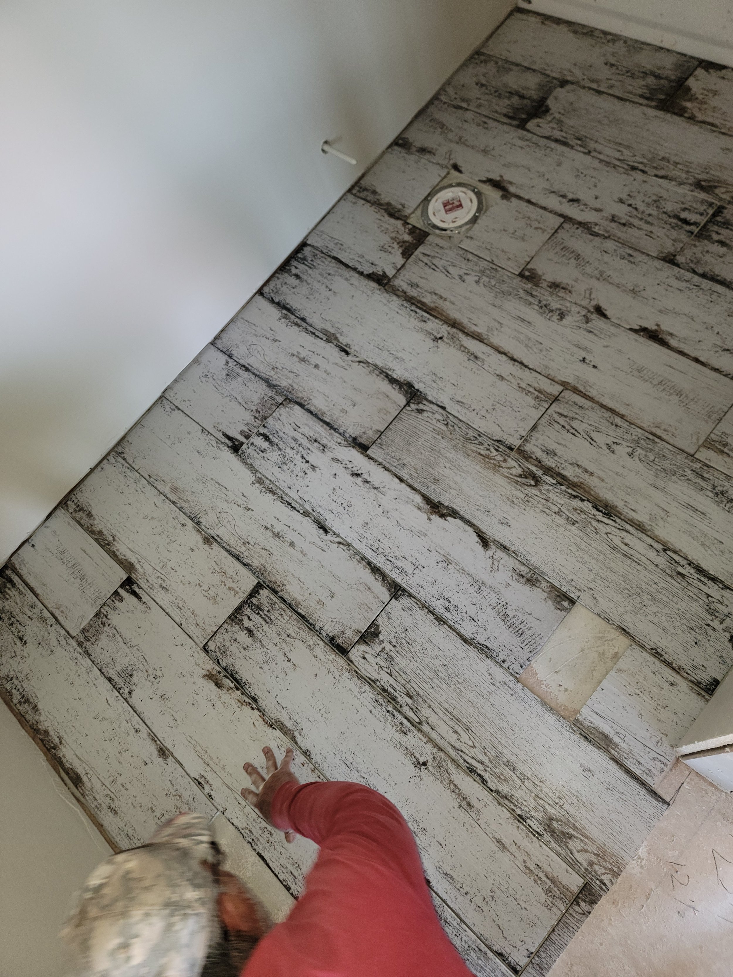 Person installing wood-look tile flooring in a room, with a dog visible at the bottom left corner.