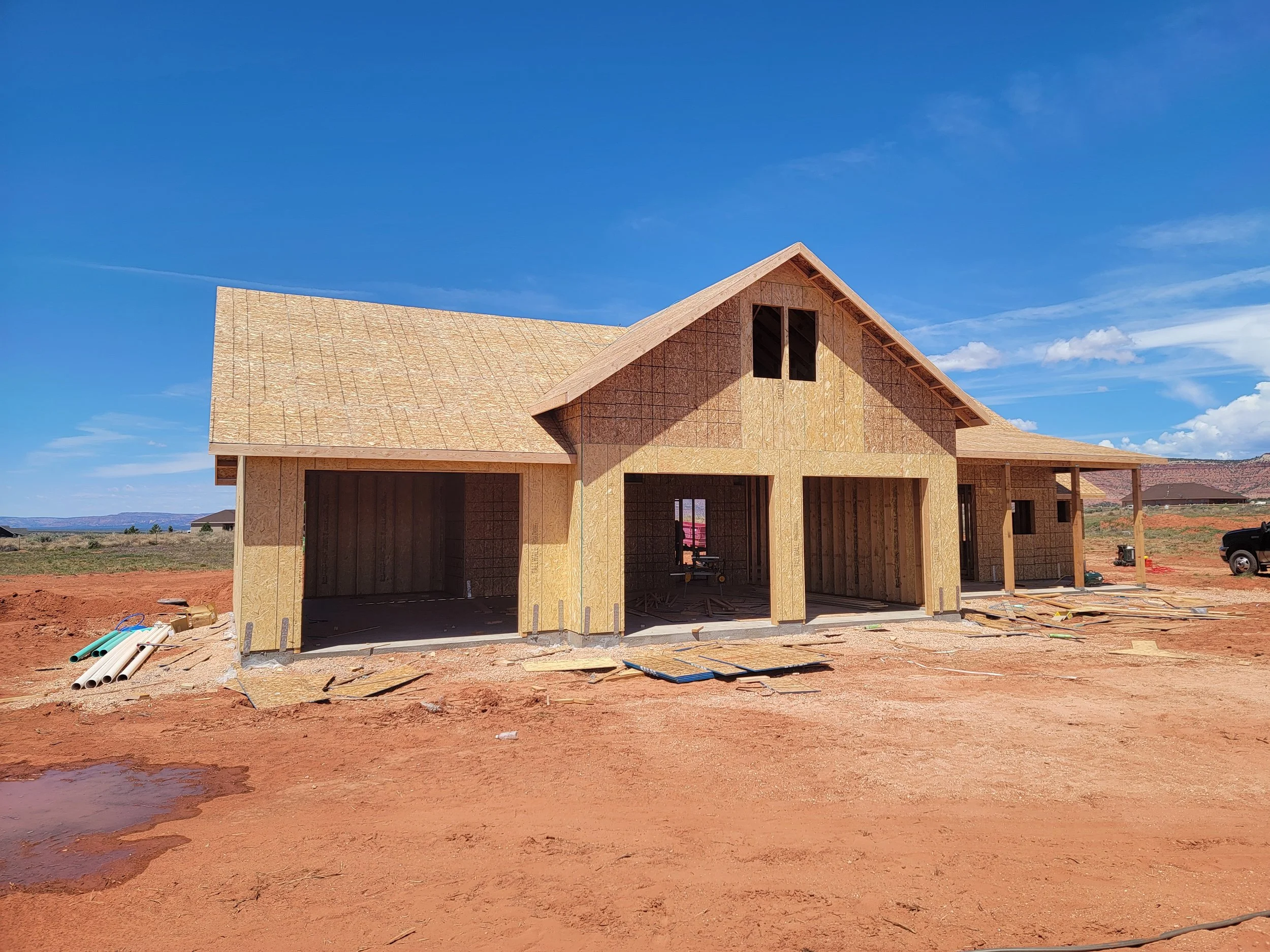 A house under construction with wooden framing, situated on a red dirt lot with construction materials nearby, under a clear blue sky.