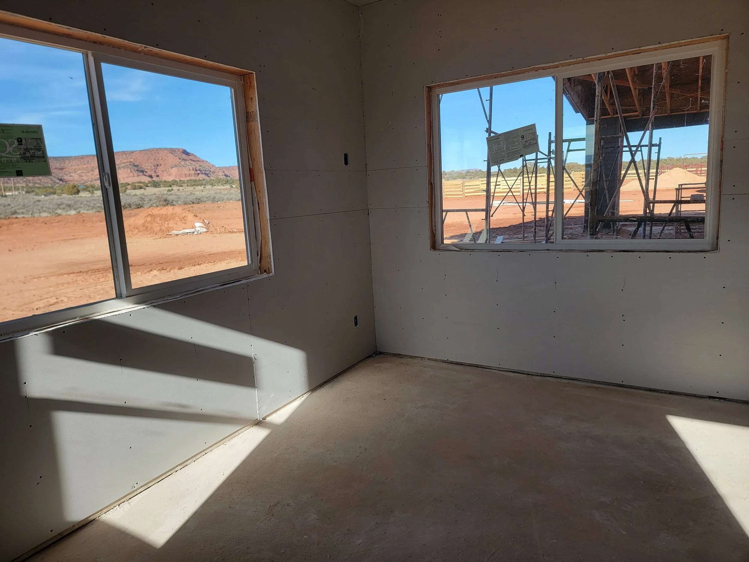 Interior view of a new building under construction with unfinished drywall and two large windows showing a desert landscape with red rocks and blue sky outside.