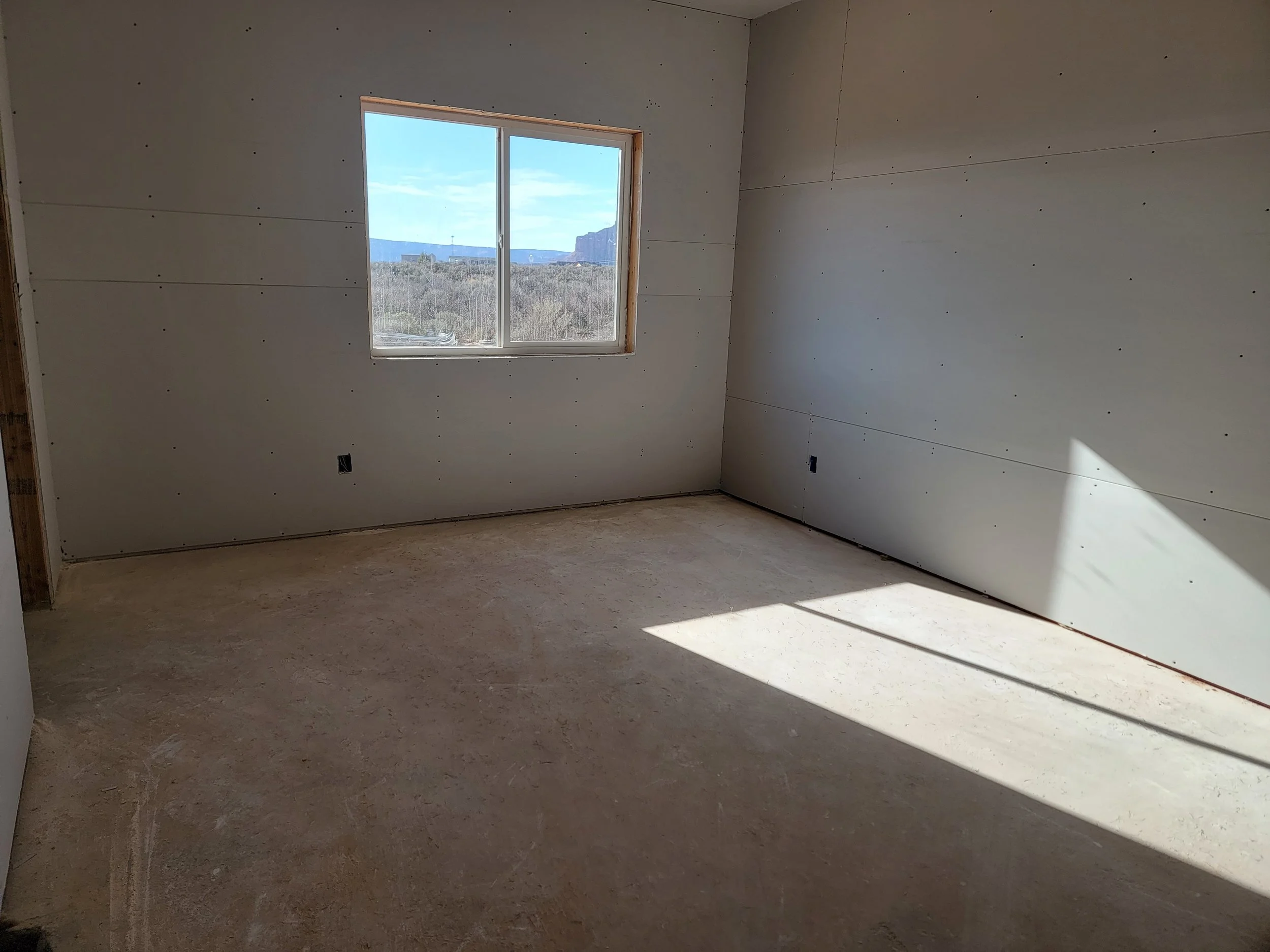 Empty room under construction with drywall on the walls, a window showing a distant landscape with mountains, and a concrete floor.