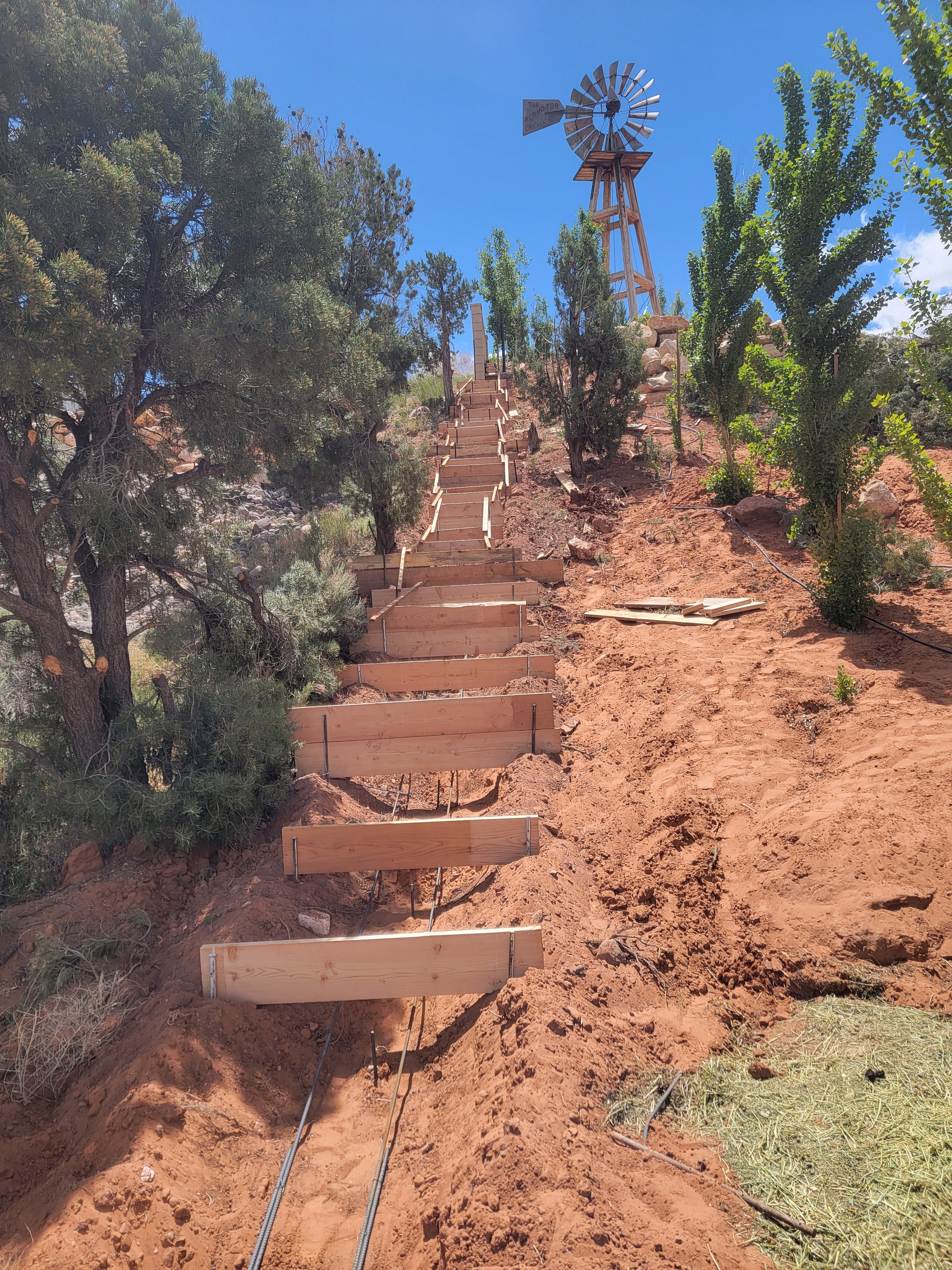 A dirt pathway with wooden forms for concrete steps, flanked by trees and bushes, leading up to an old windmill with a weather vane on top under a clear blue sky.