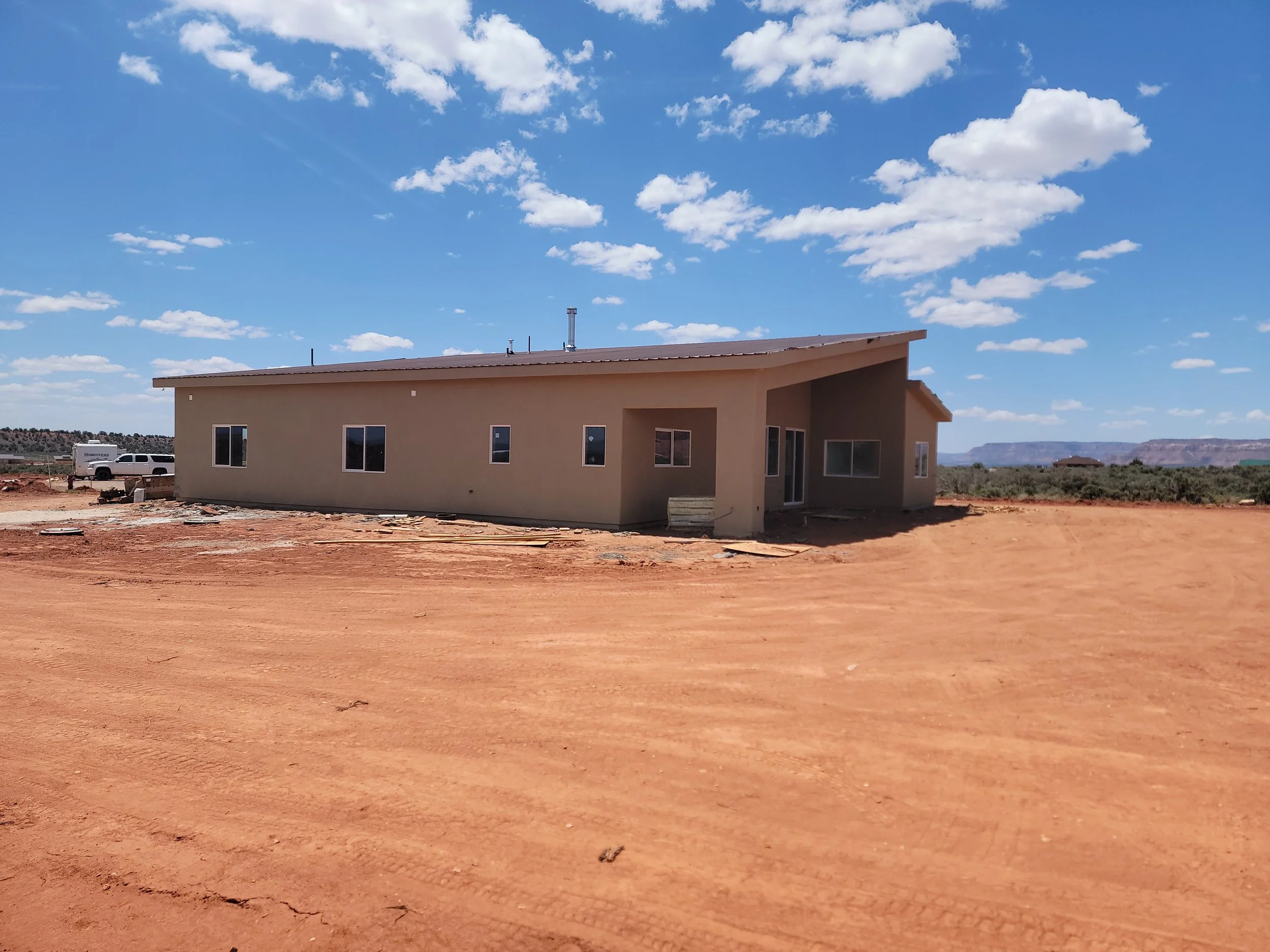 A house under construction in a rural area with a dirt ground and clear blue sky with scattered clouds.