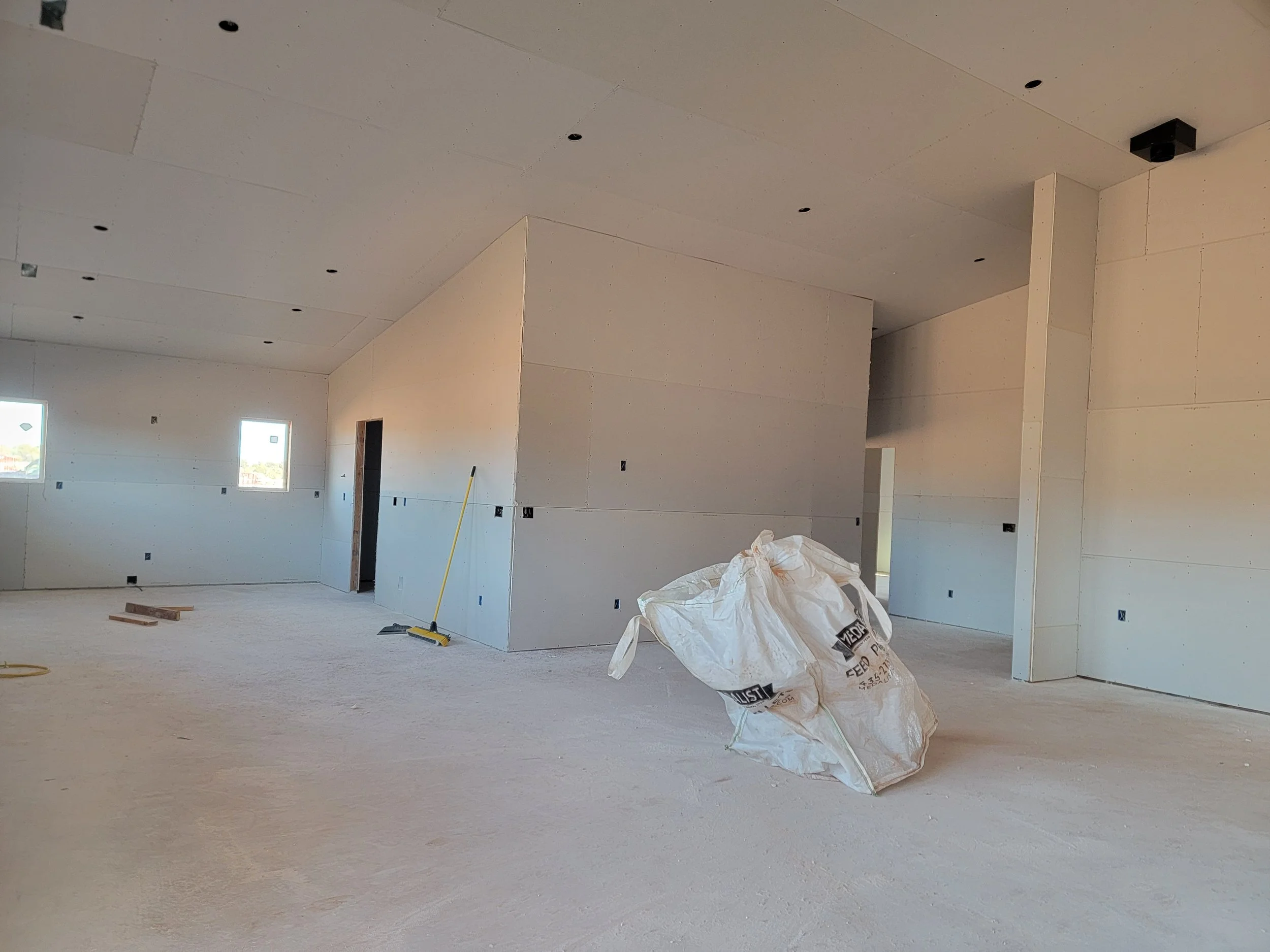 Interior of a building under construction with unfinished walls and ceiling, construction materials, and tools visible, including a large bag of construction feed and a broom.