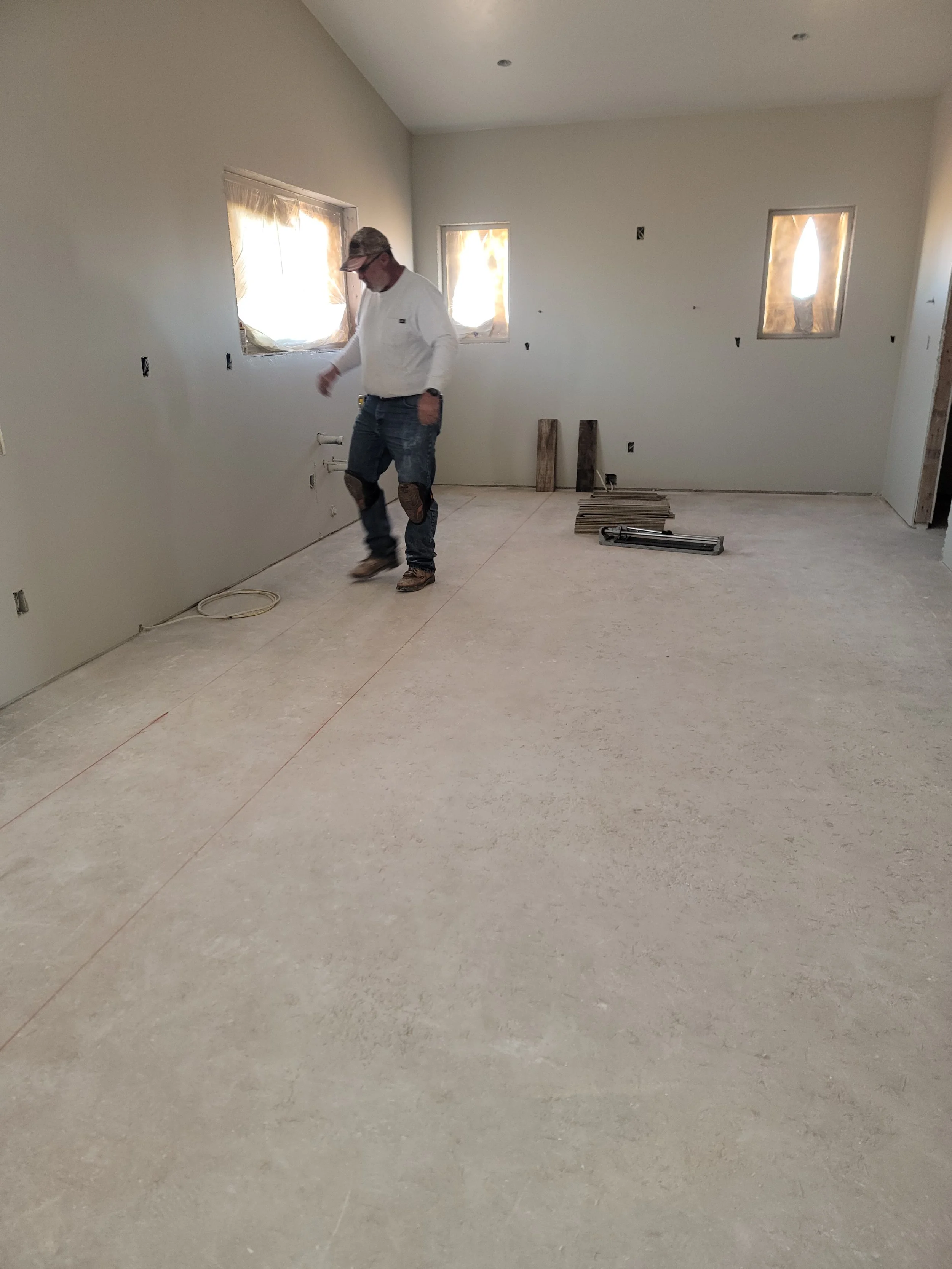 Man working on a construction or renovation project in an unfinished room with three small windows, a drywall wall, and tools on the floor.