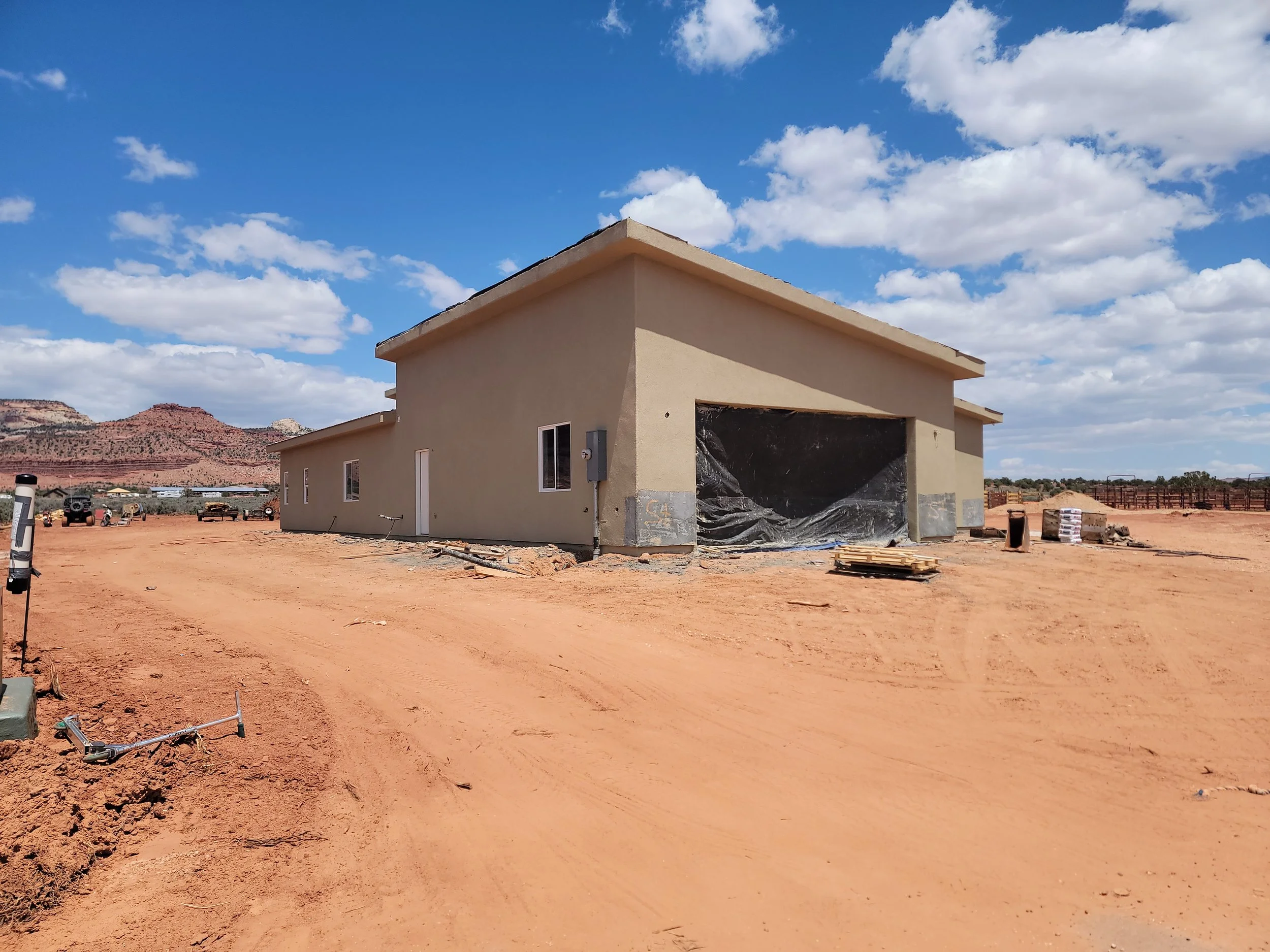 Under construction house with a beige exterior, set in a desert landscape with red soil, mountains, and a blue sky with clouds.