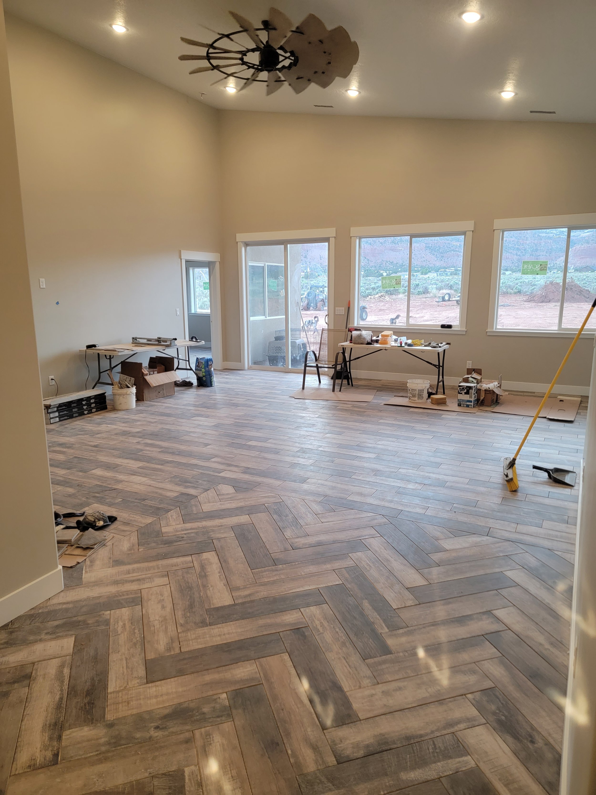 Living room under renovation with new wood-look tile flooring, construction tools, tables, and large windows showing outdoor landscape.