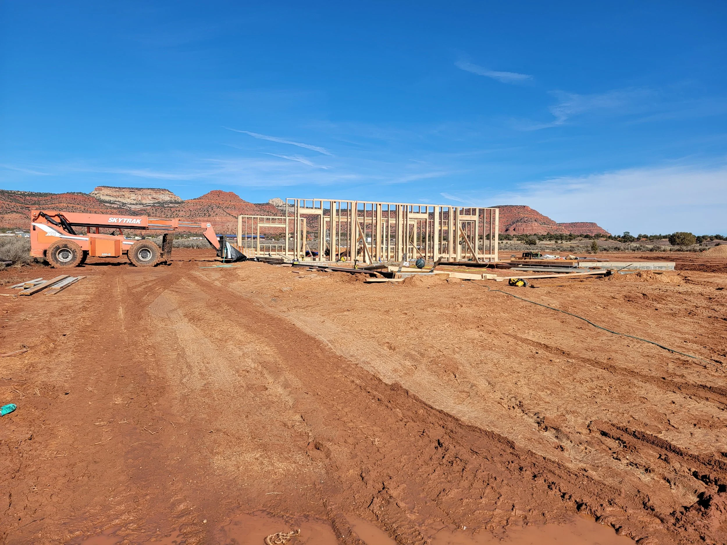 Construction site with wooden framing and construction equipment in a desert landscape with hills in the background.