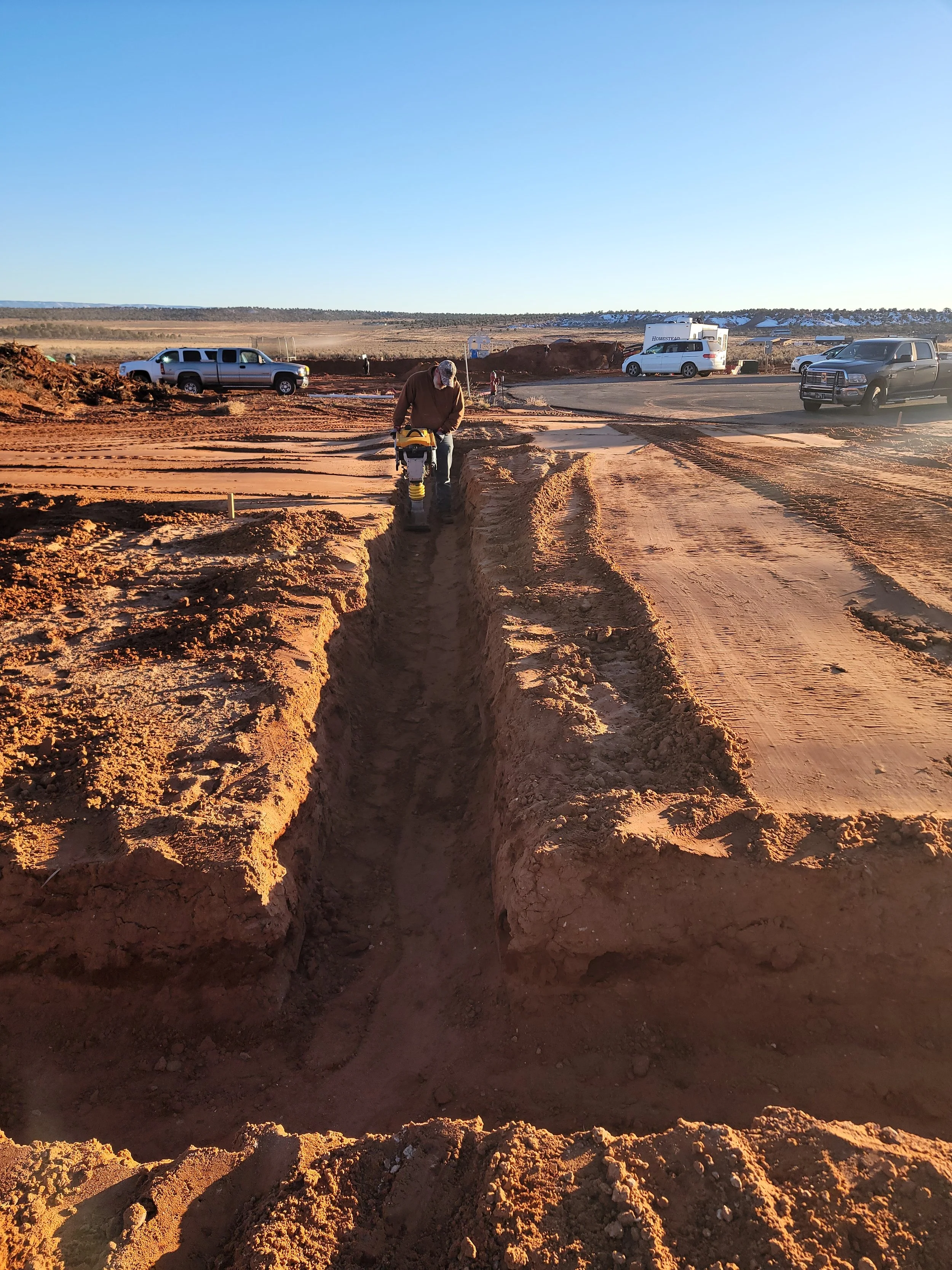 A construction worker operating a compaction device in a dirt trench at a construction site during daylight. Several parked vehicles, including trucks and an RV, are visible in the background under a clear blue sky.