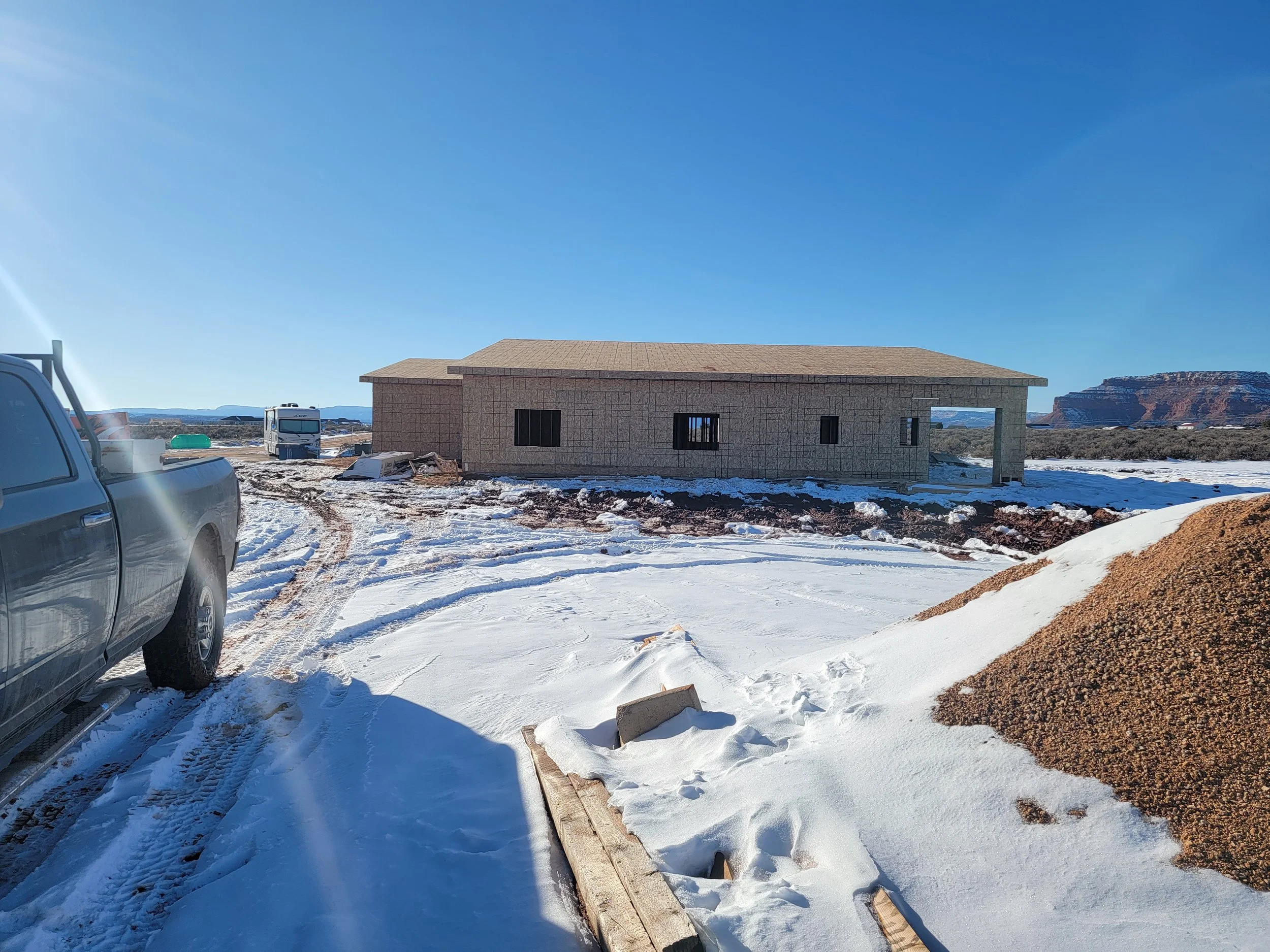 Construction site with a single-story house, construction vehicles, and snow on the ground in a rural area with mountains in the background.