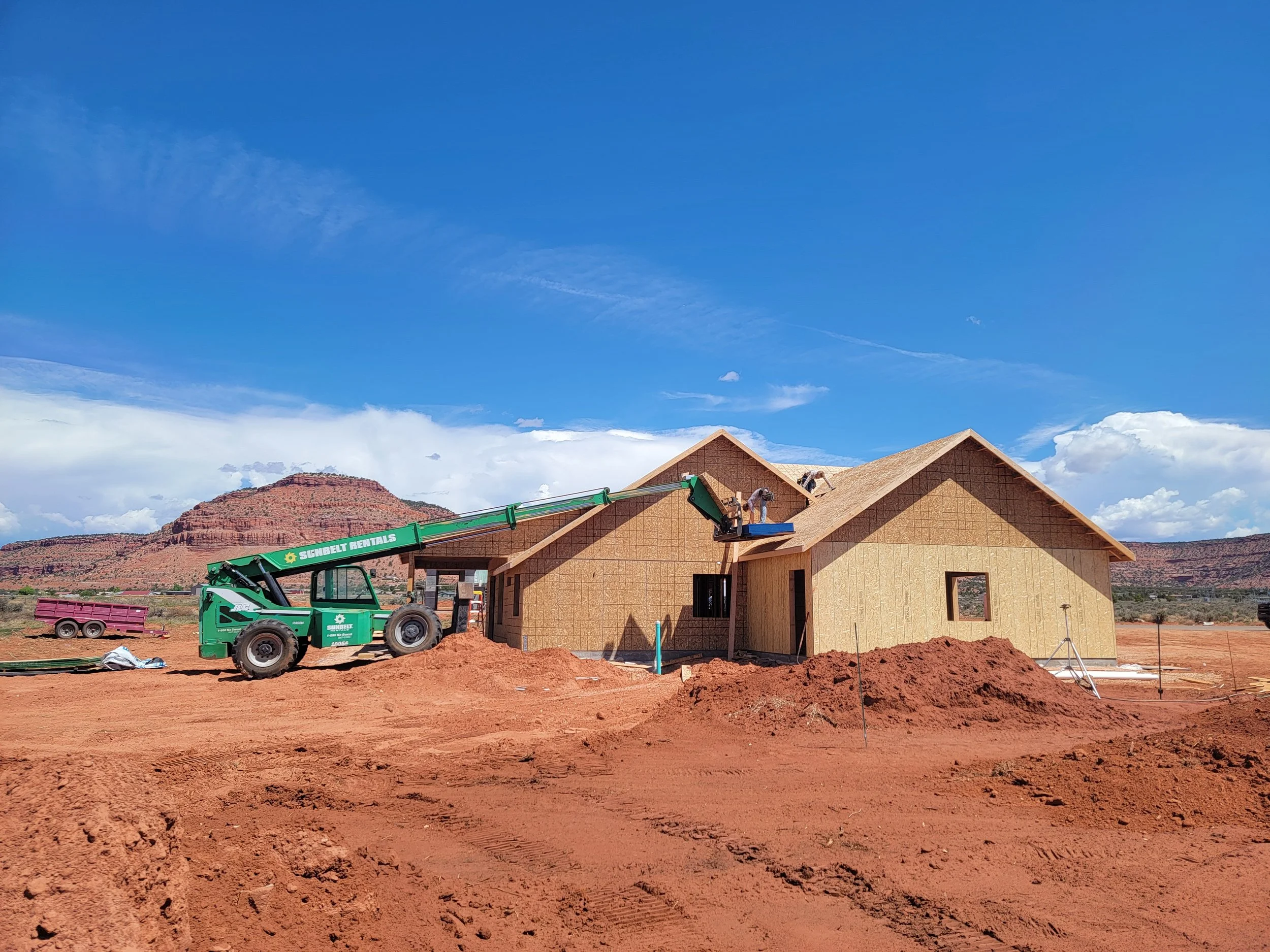 Construction site with a house under construction, showing a worker on the roof, a green crane, and a large pile of reddish dirt in the foreground. The background features a blue sky with some clouds and a mountain range.
