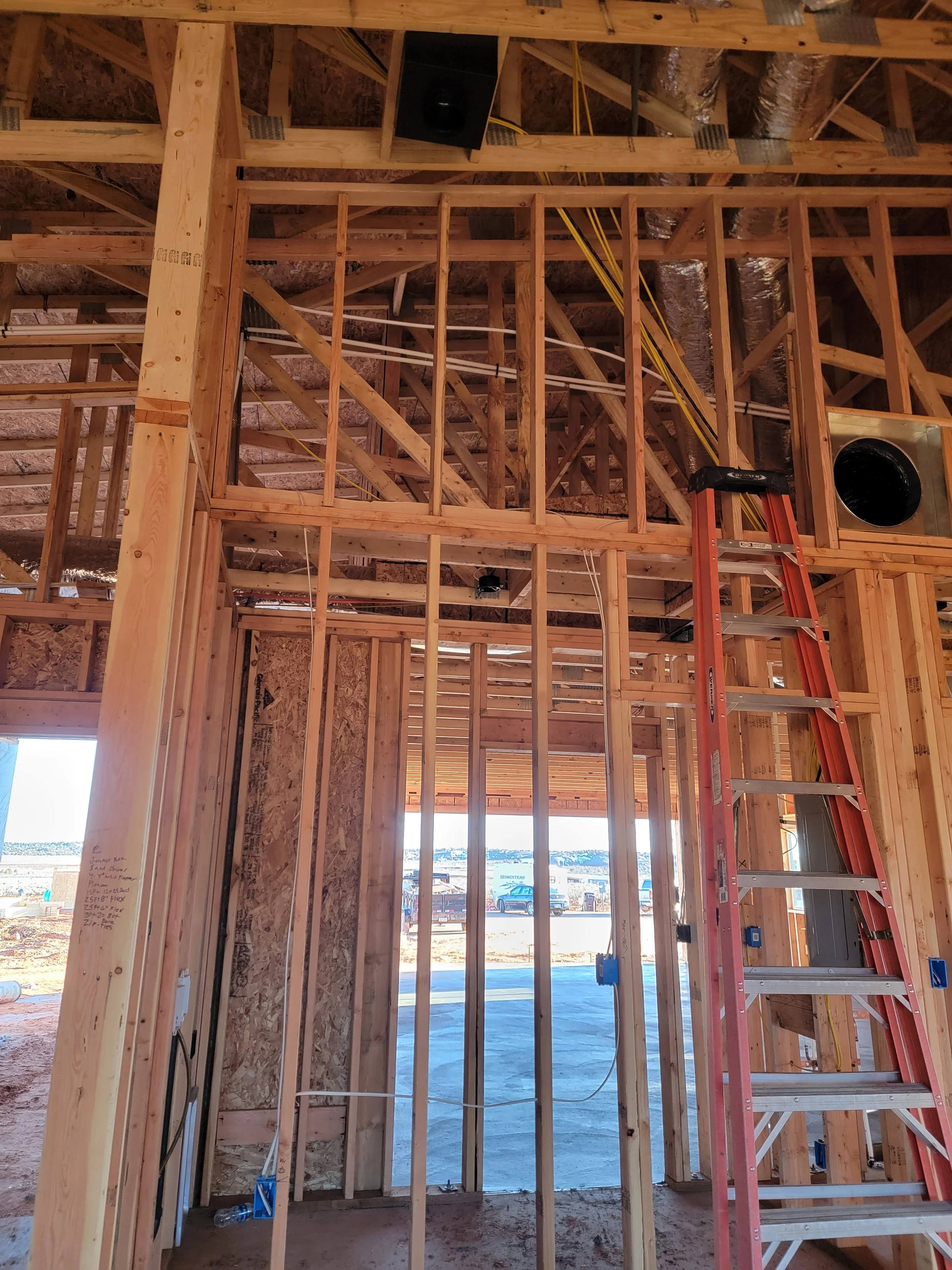 The interior of a house under construction showing a wooden framing structure, an orange ladder, and an opening in the wall with a view outside.