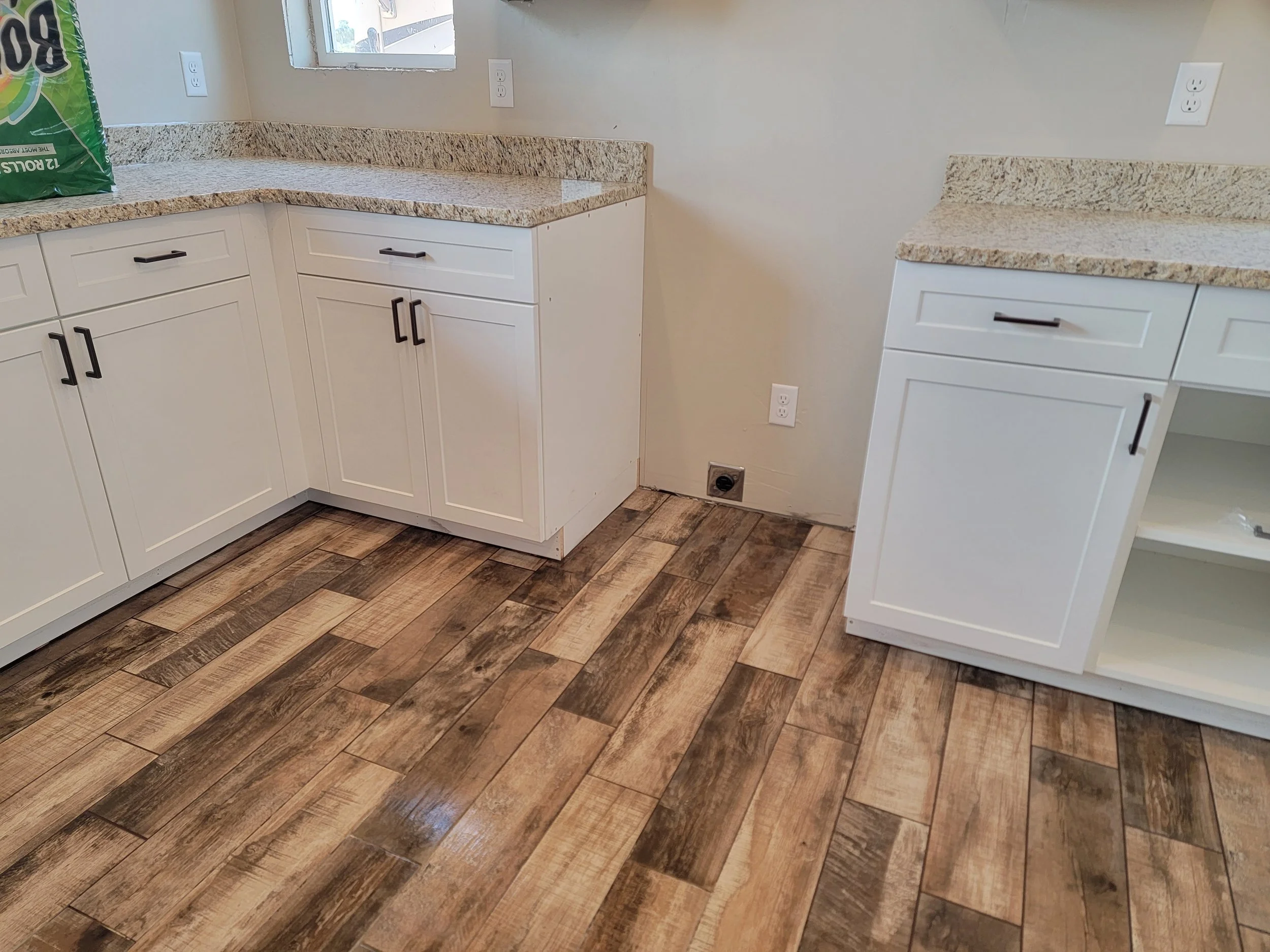 Kitchen with white cabinets, granite countertops, and wood-look tile flooring. A window above the countertop, and electrical outlets on the wall.