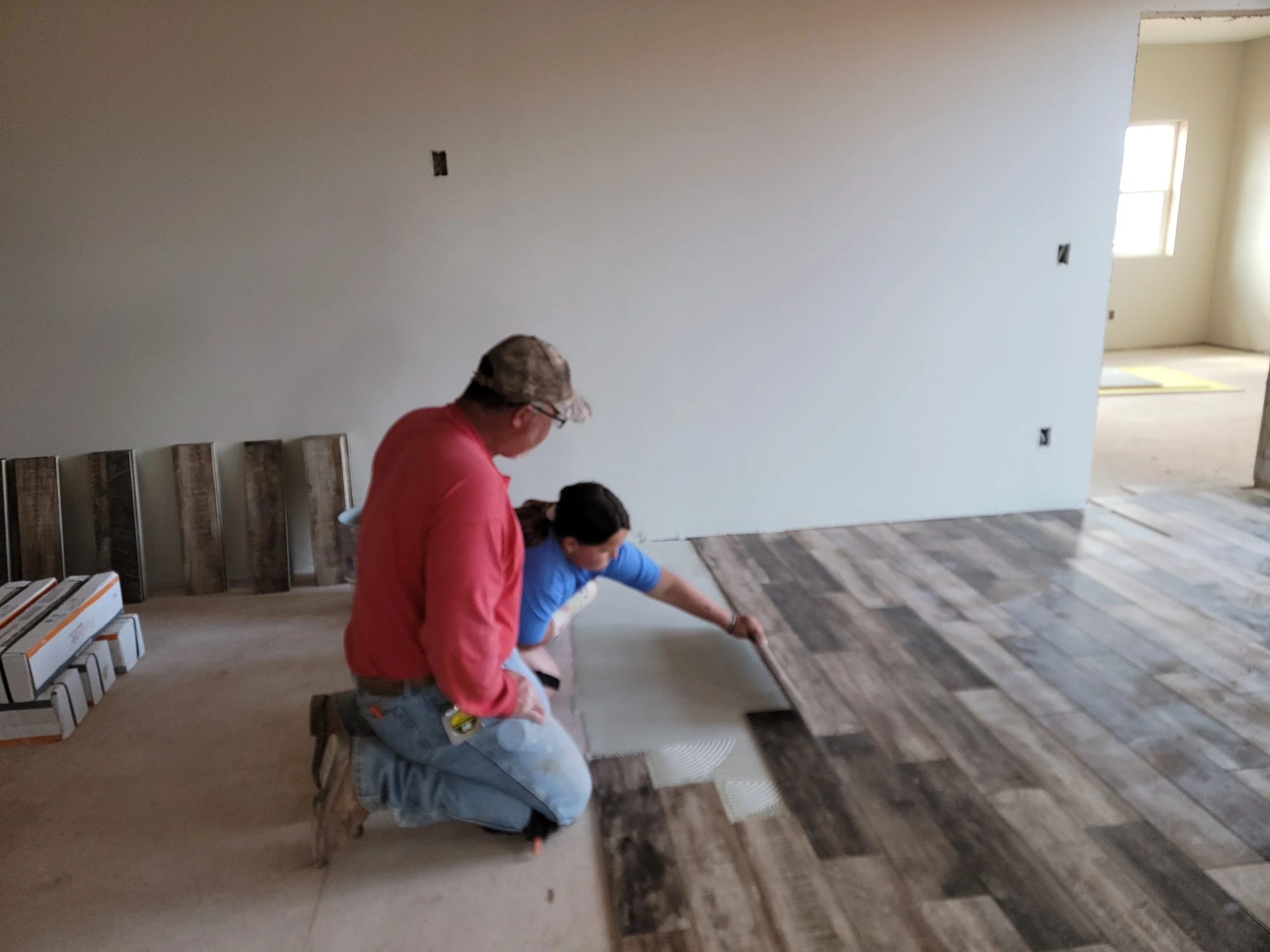 Two people working on a flooring project inside a house, with one person kneeling and applying flooring while the other person stands nearby.