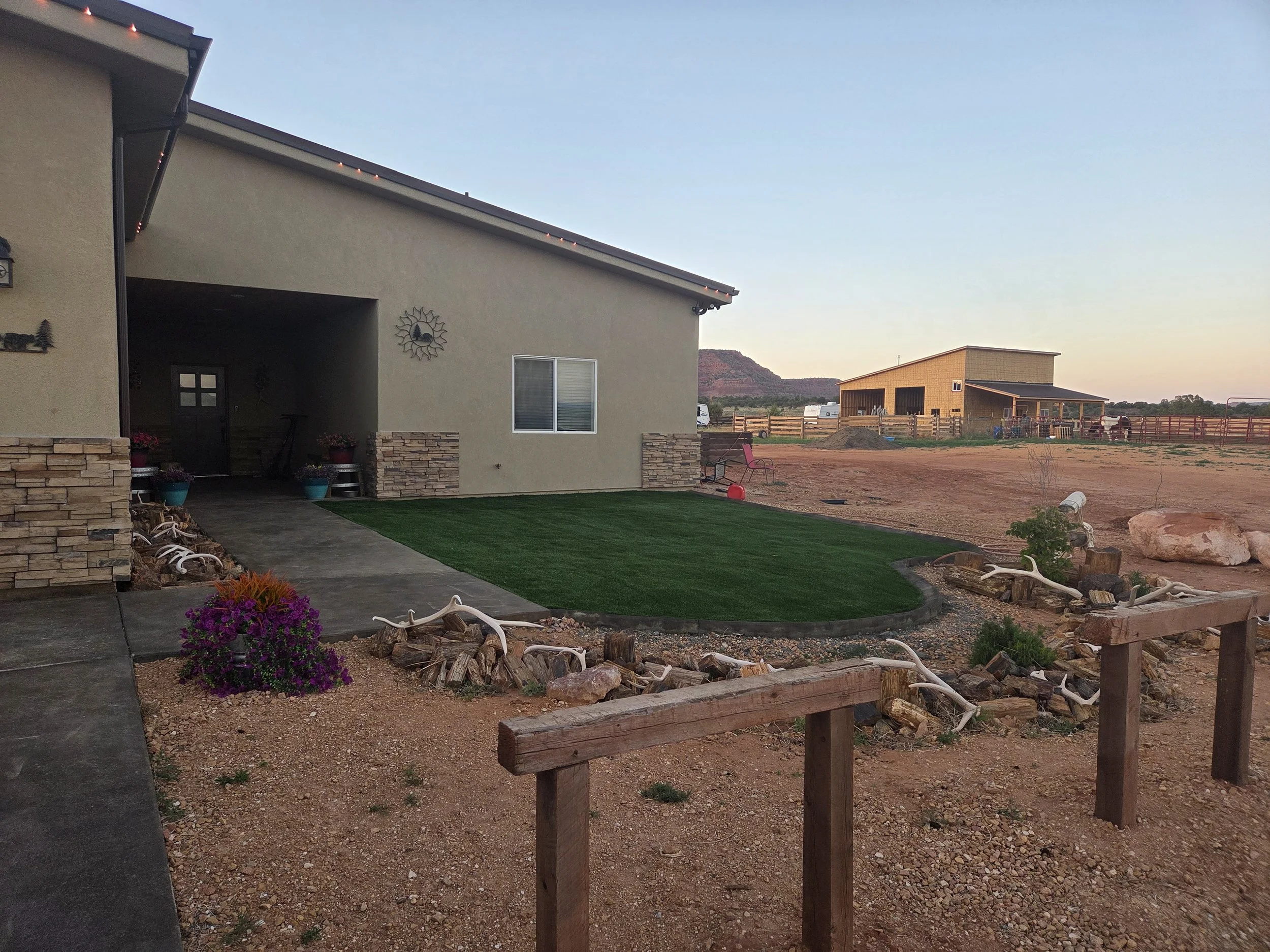 Photo of the backyard of a house during late afternoon or early evening with a patch of artificial grass, a wooden fence, a pink chair, potted plants, and a mountain with reddish slopes in the background.