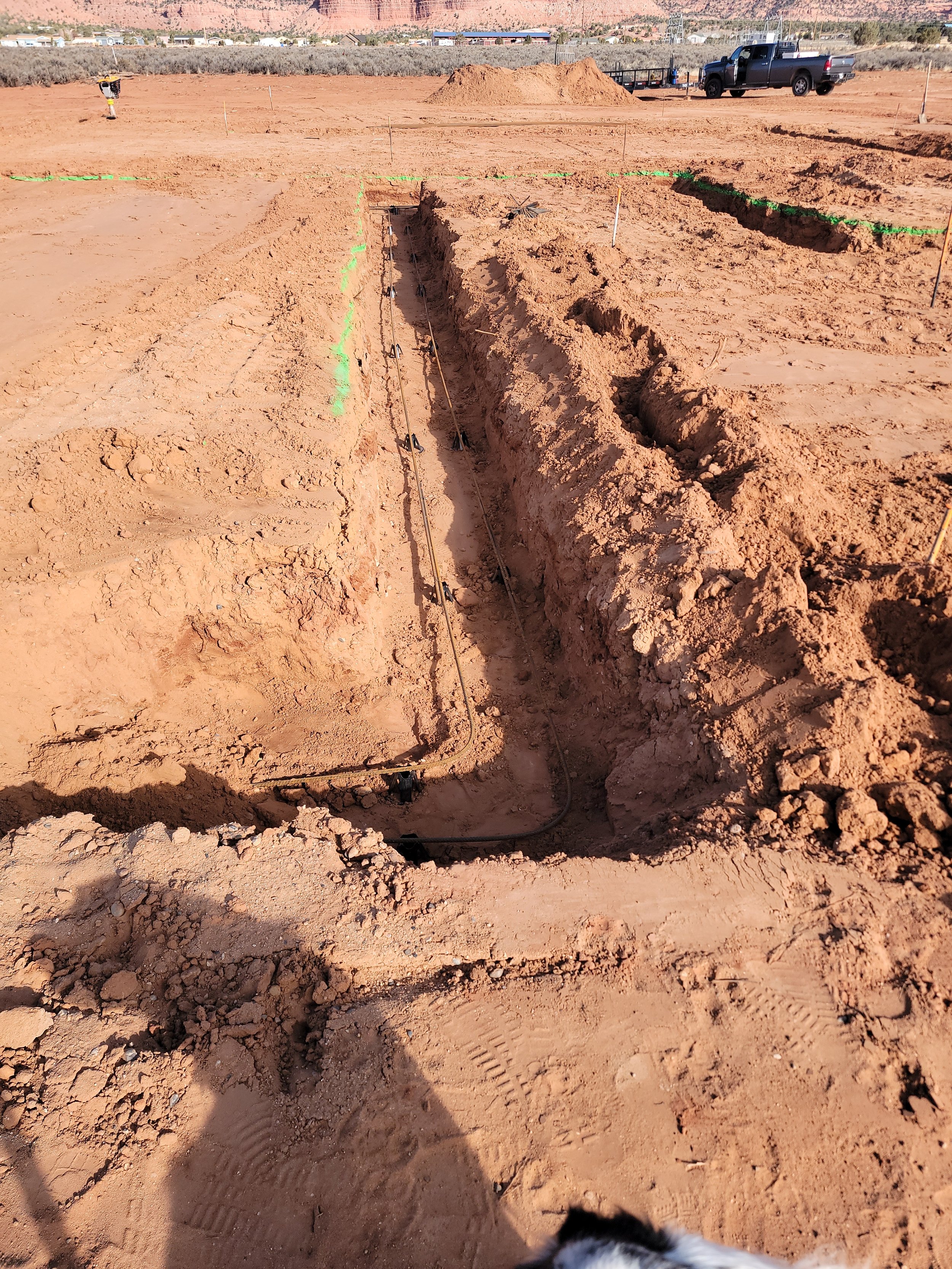 Construction site with trenches and wiring in red dirt, equipment and a truck in the background, and a small dog in the lower right corner.