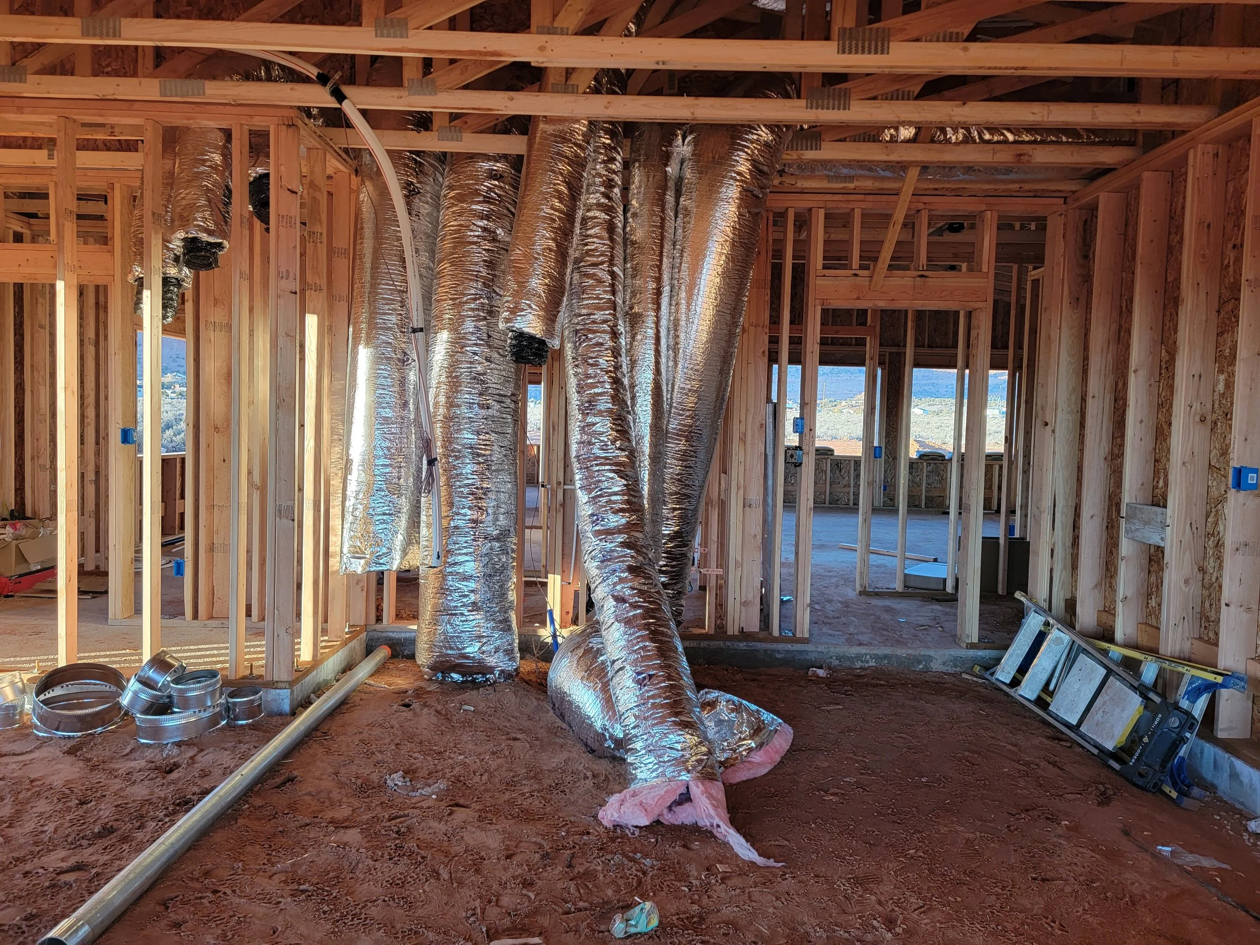 Construction site with exposed wooden framing and large silver flexible ducts, insulation on the floor, and construction tools.