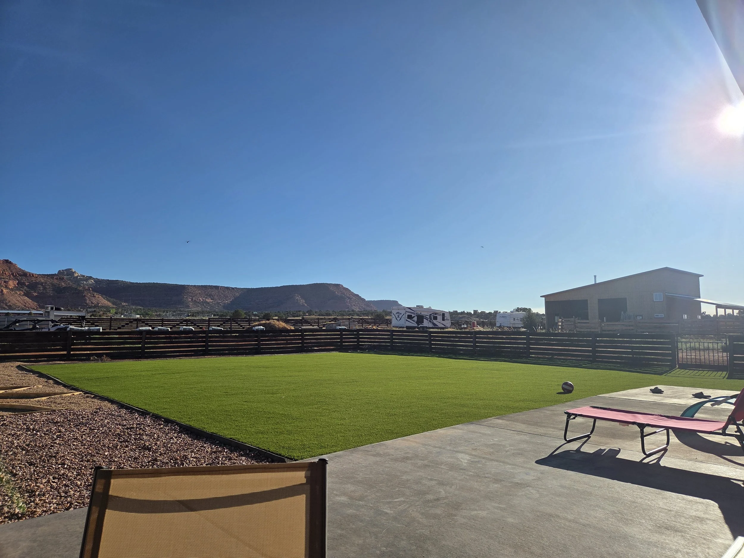A backyard with a trampoline, a pink lounge chair, a small soccer ball, and a green artificial turf area under a clear blue sky with a bright sun.