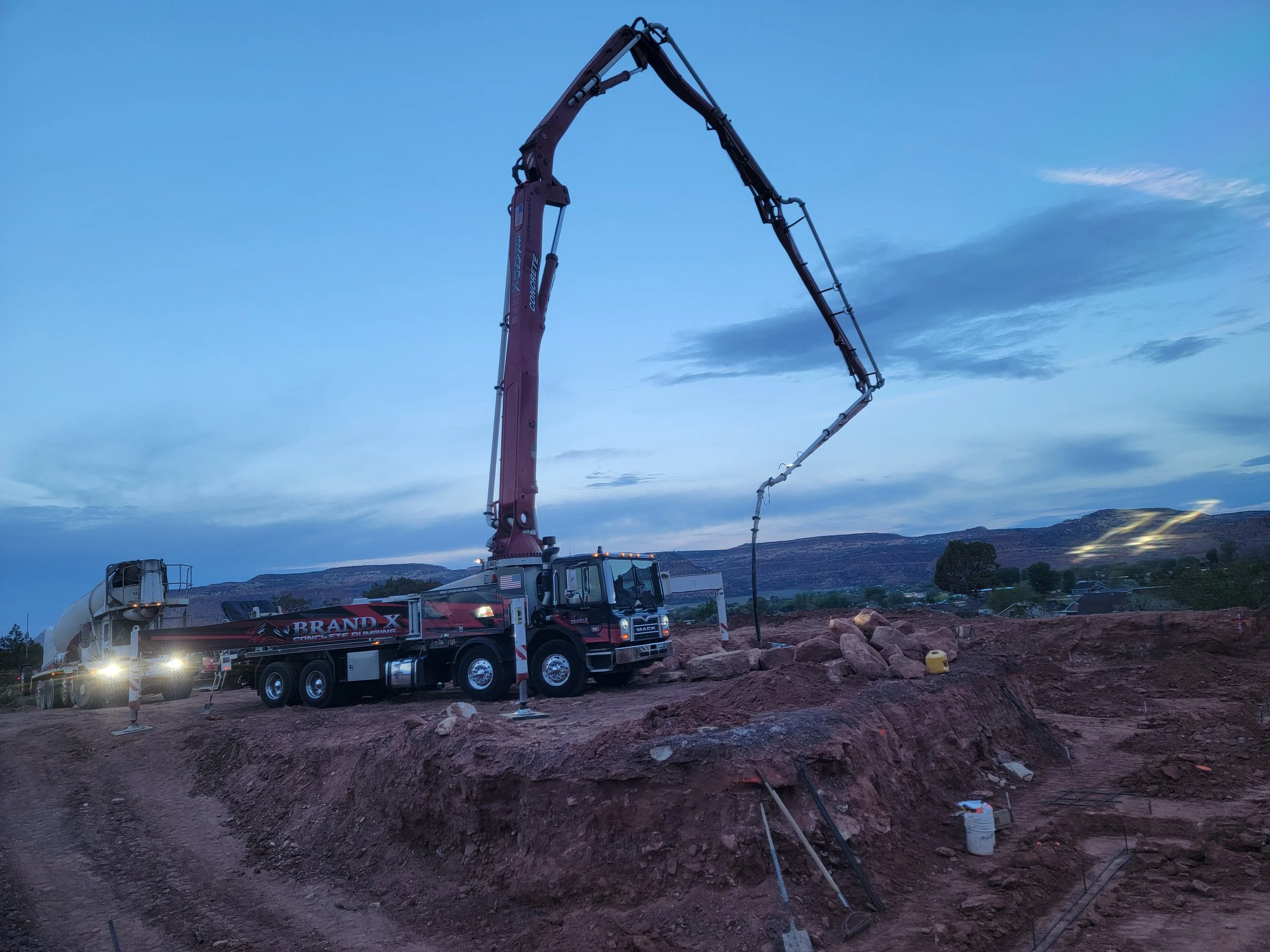 A large concrete pump truck with a long extended boom is operating on a construction site during dusk, with mountains and a cloudy sky in the background.