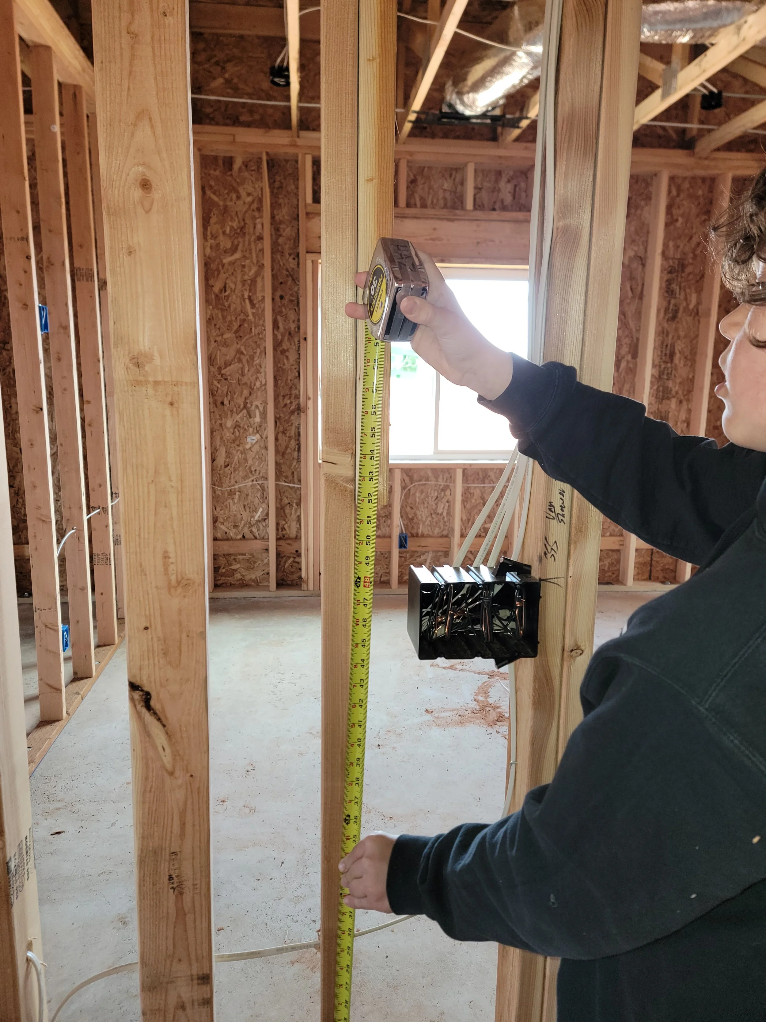 A person measures a wooden beam with a tape measure within a house under construction, showing the interior framing and exposed wooden studs.