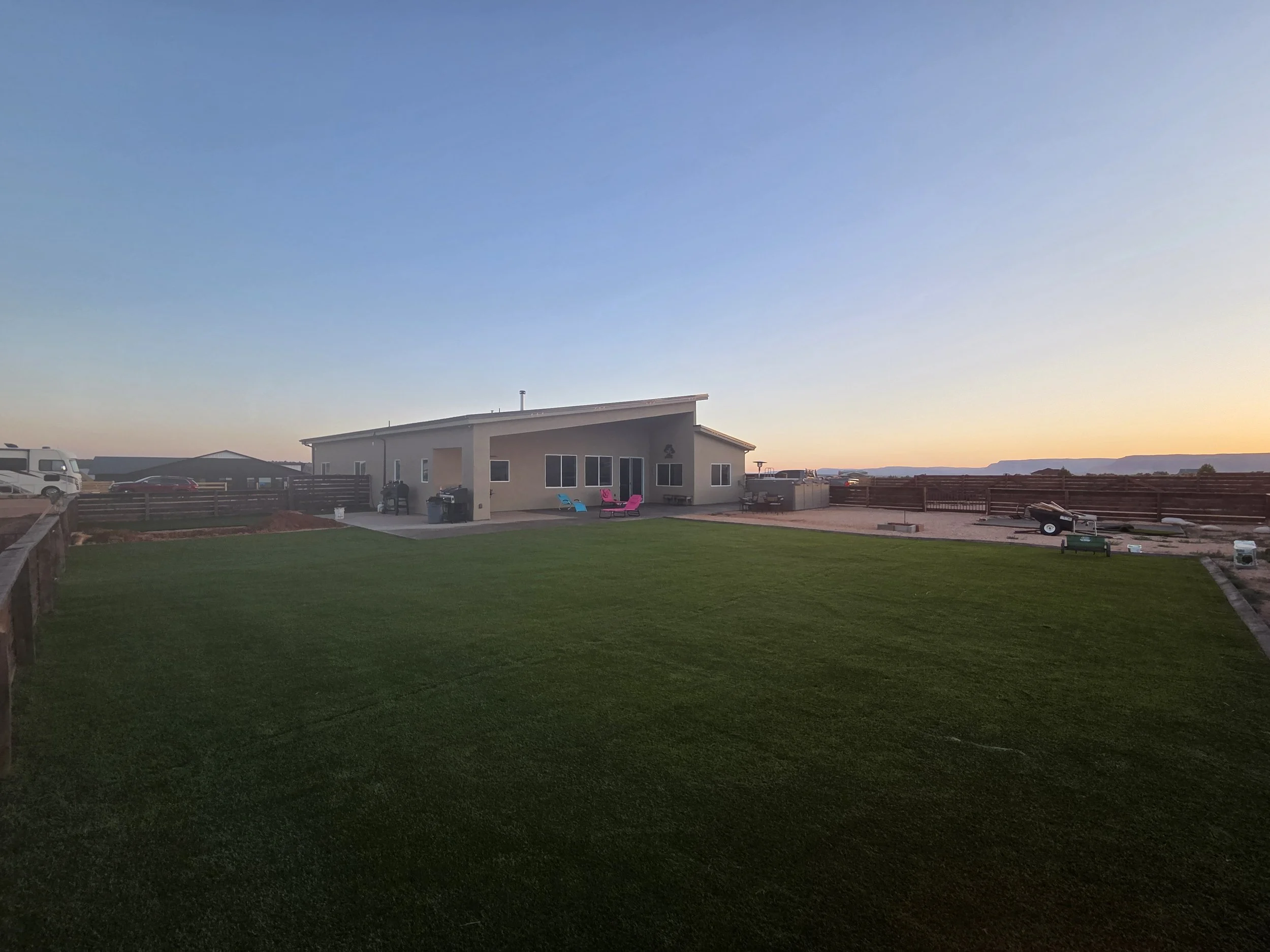 View of a backyard during sunset with a modern house, outdoor seating, and a green lawn.