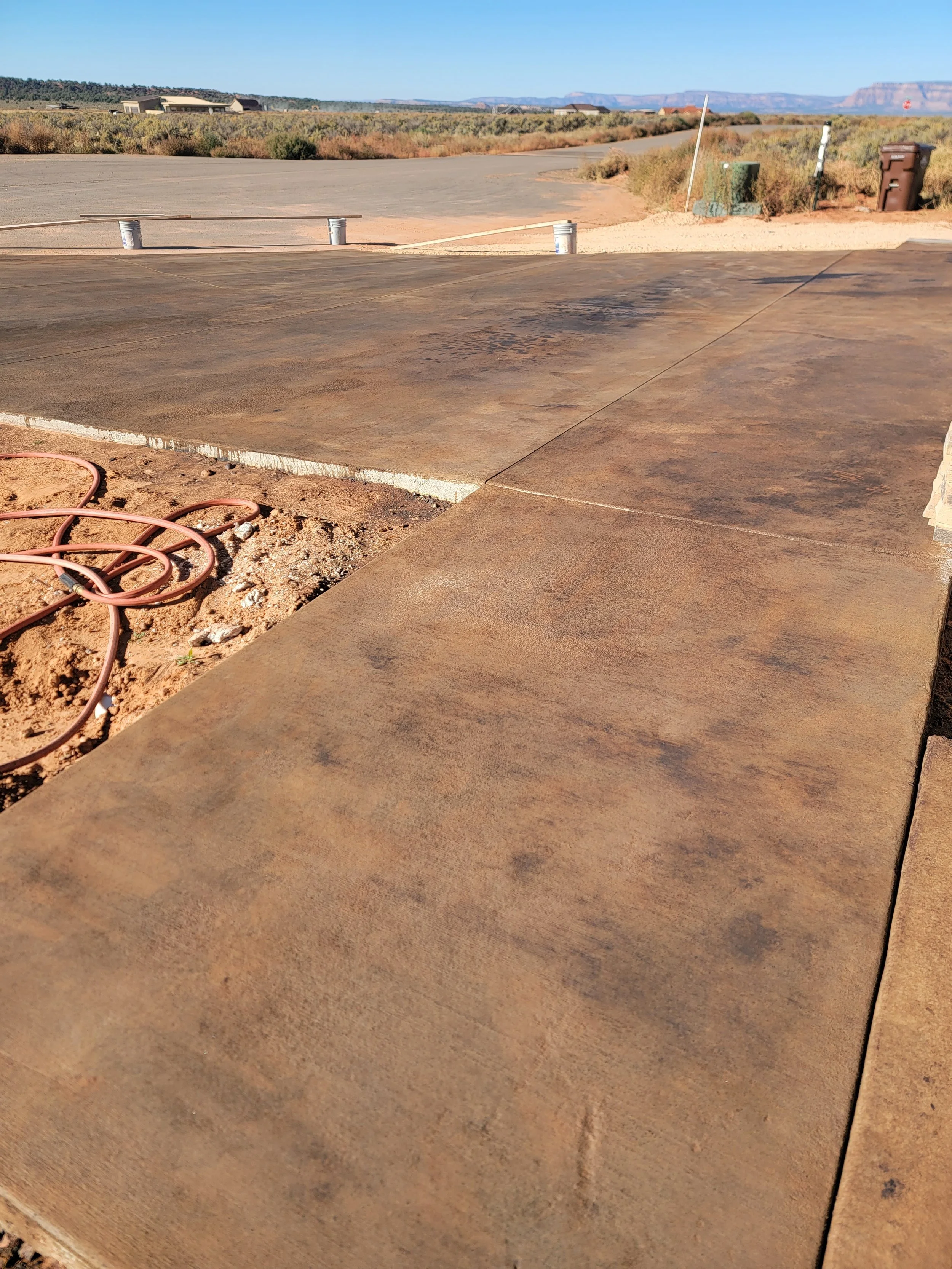 A newly laid concrete patio with visible expansion joints, garden hoses, and a remote desert landscape in the background.