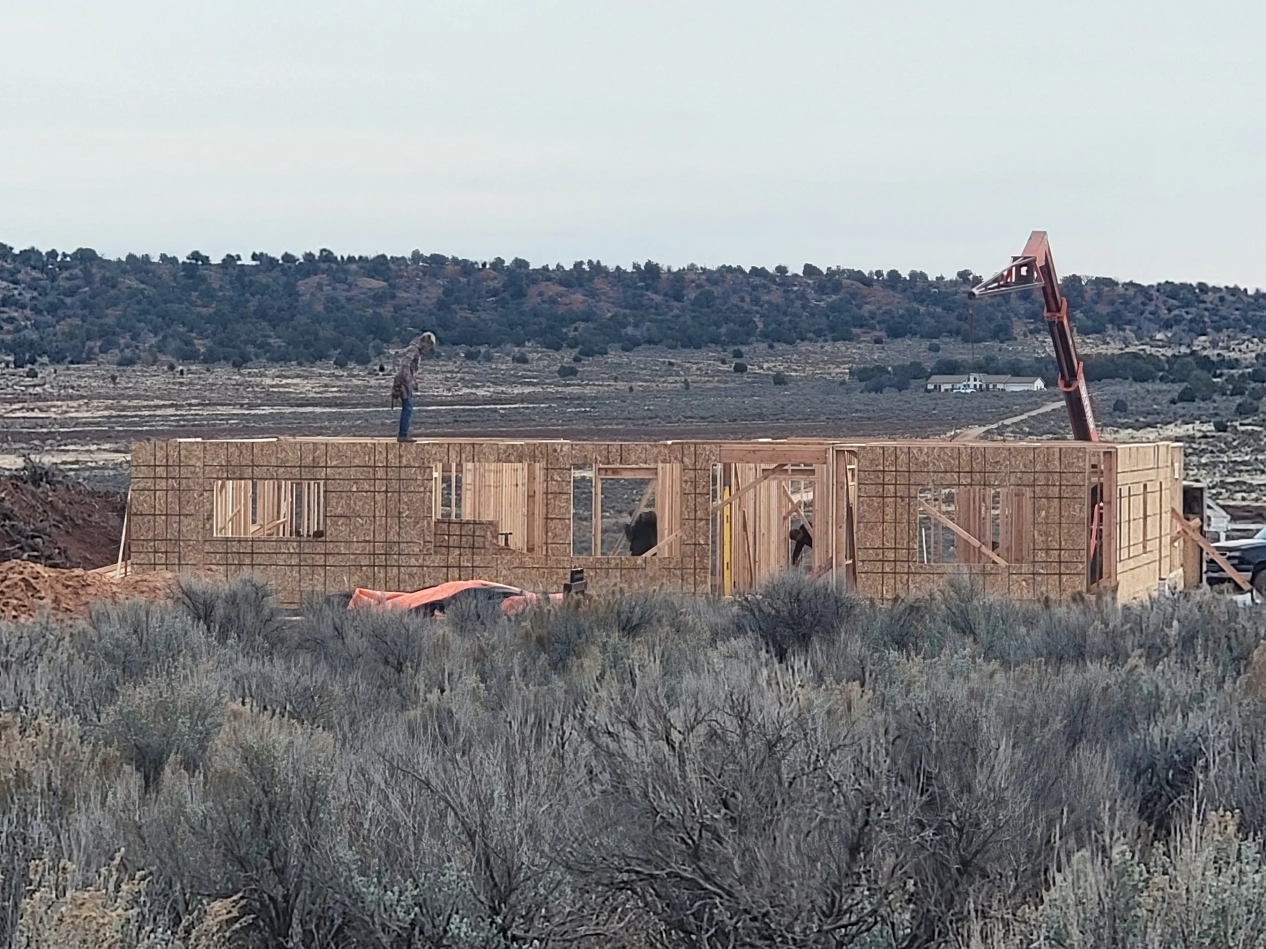 A construction site in a rural area with a wooden framed building under construction, a person on the roof, and a large red crane nearby, with hills and sparse vegetation in the background.