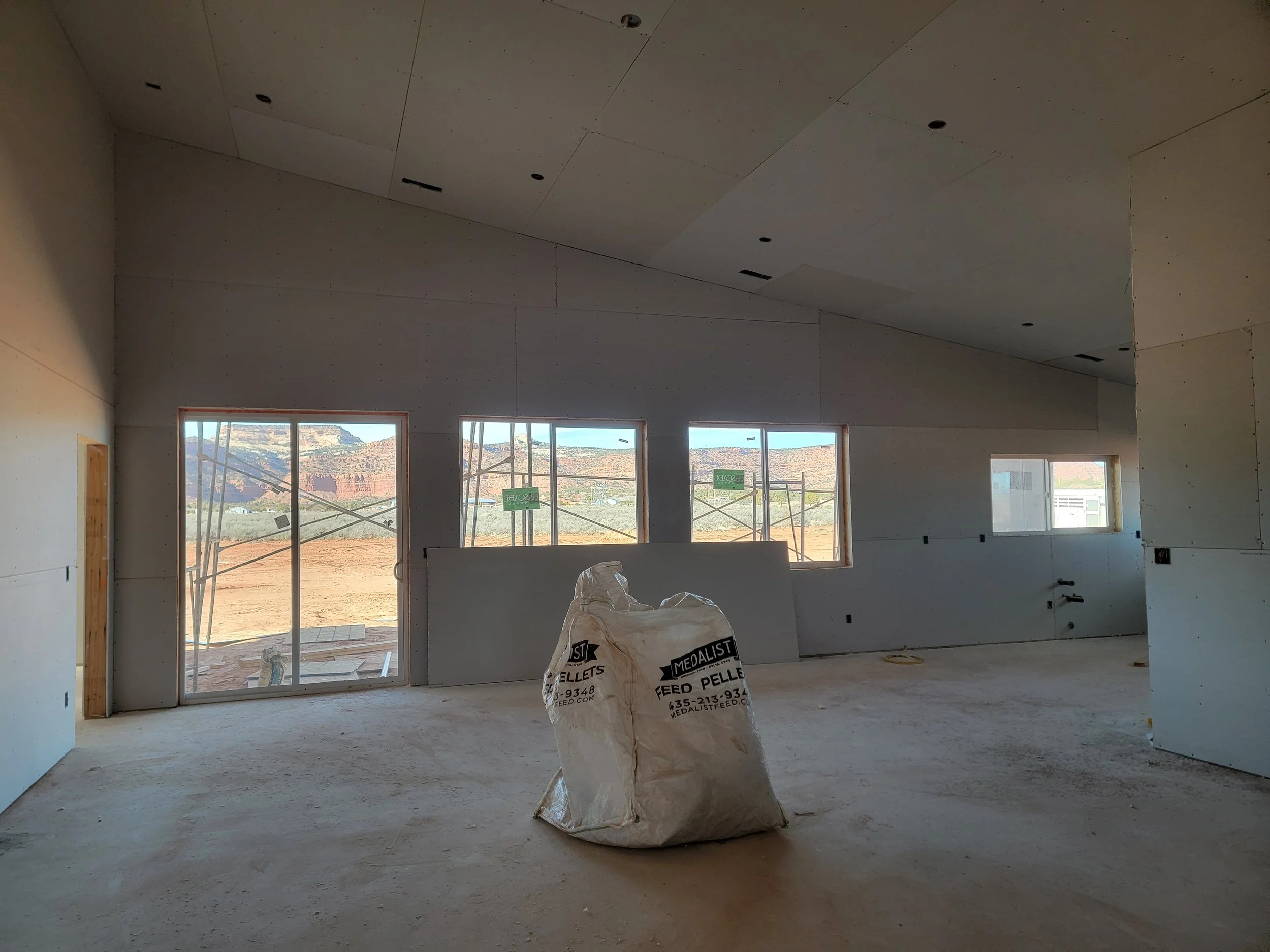 Interior of a building under construction with unfinished drywall ceiling and walls, large windows showing a desert landscape, and construction materials on the floor.