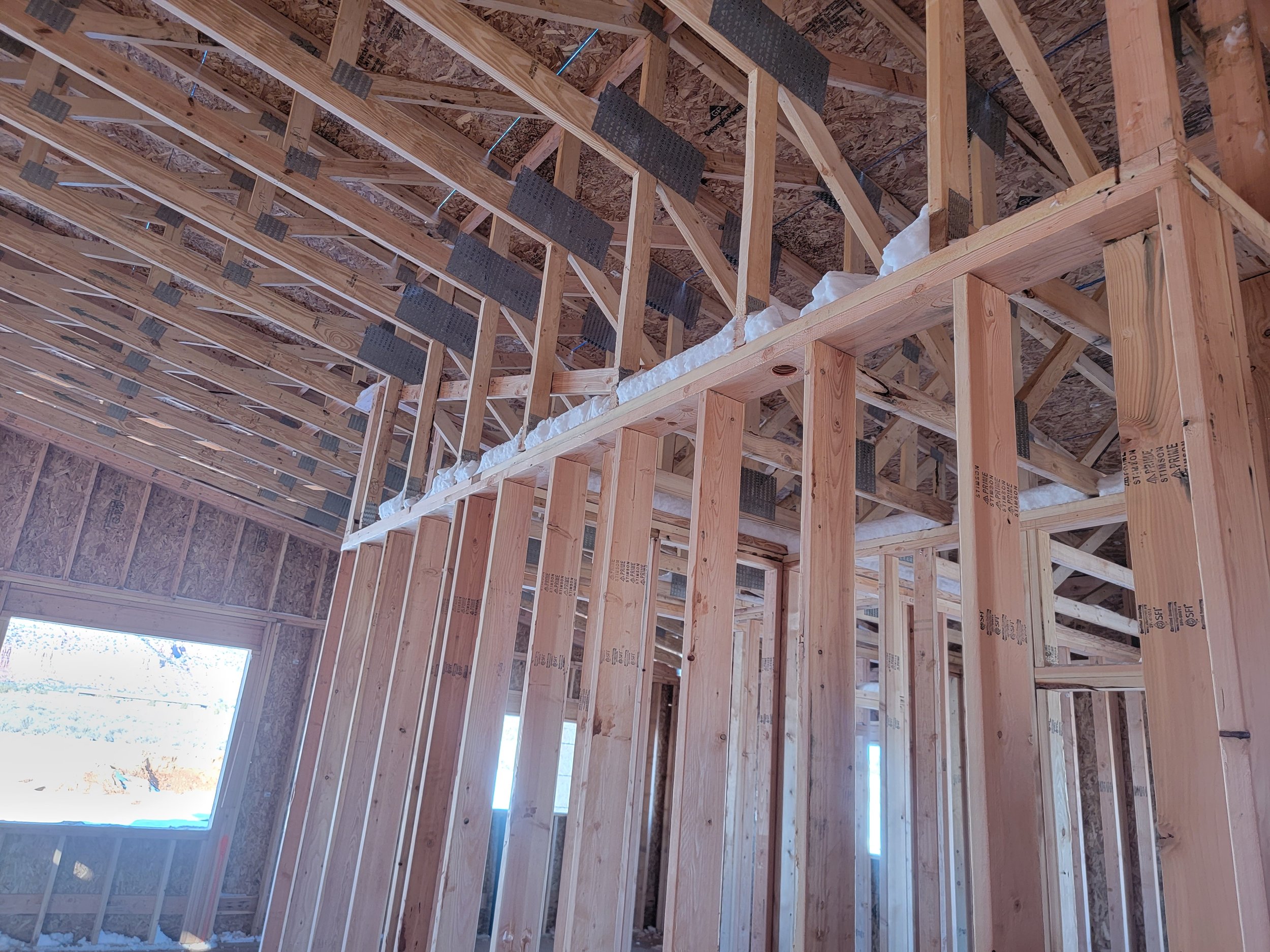 The interior wooden framework of a building under construction, including vertical studs, horizontal beams, and roofing trusses, with windows allowing natural light inside.