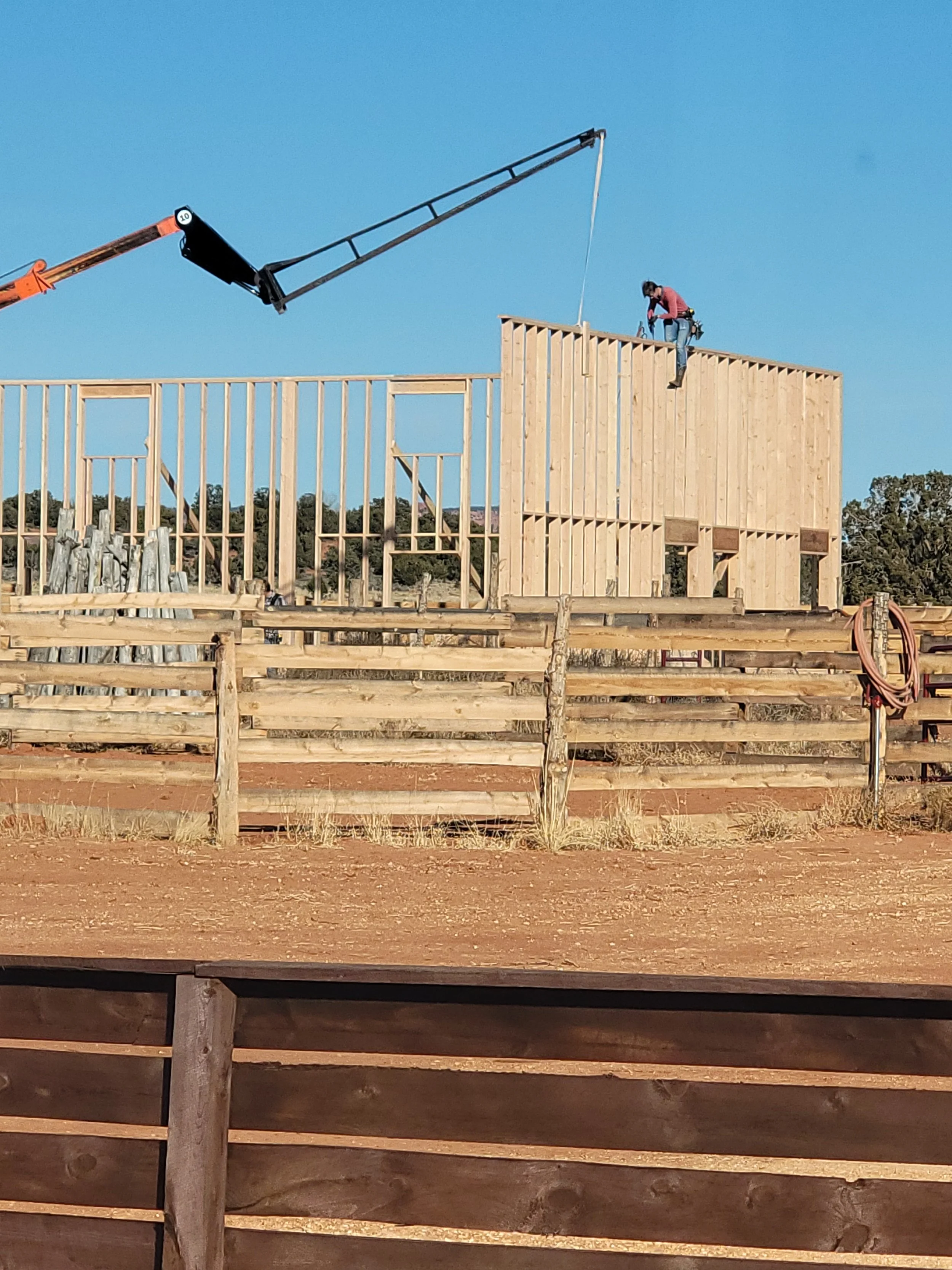 A construction site with a wooden structure in progress and a worker on top, using a crane to lift materials against a blue sky with trees in the background.