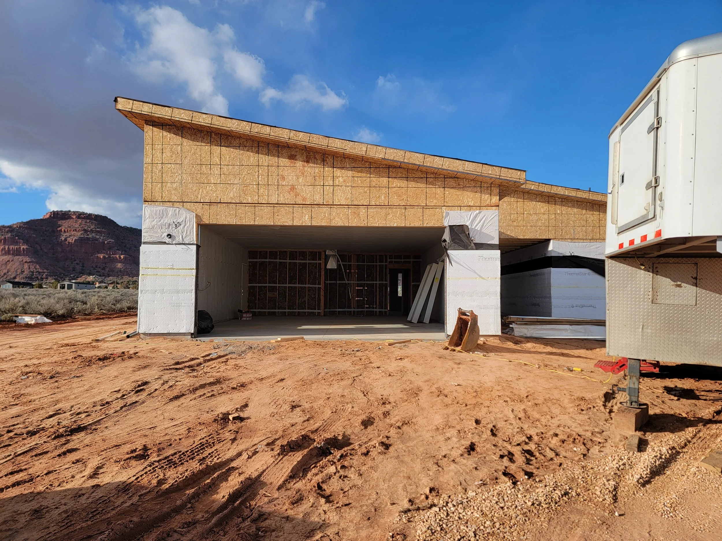 Construction site with a partially built wooden house and a white trailer in a desert area, mountains in the background.