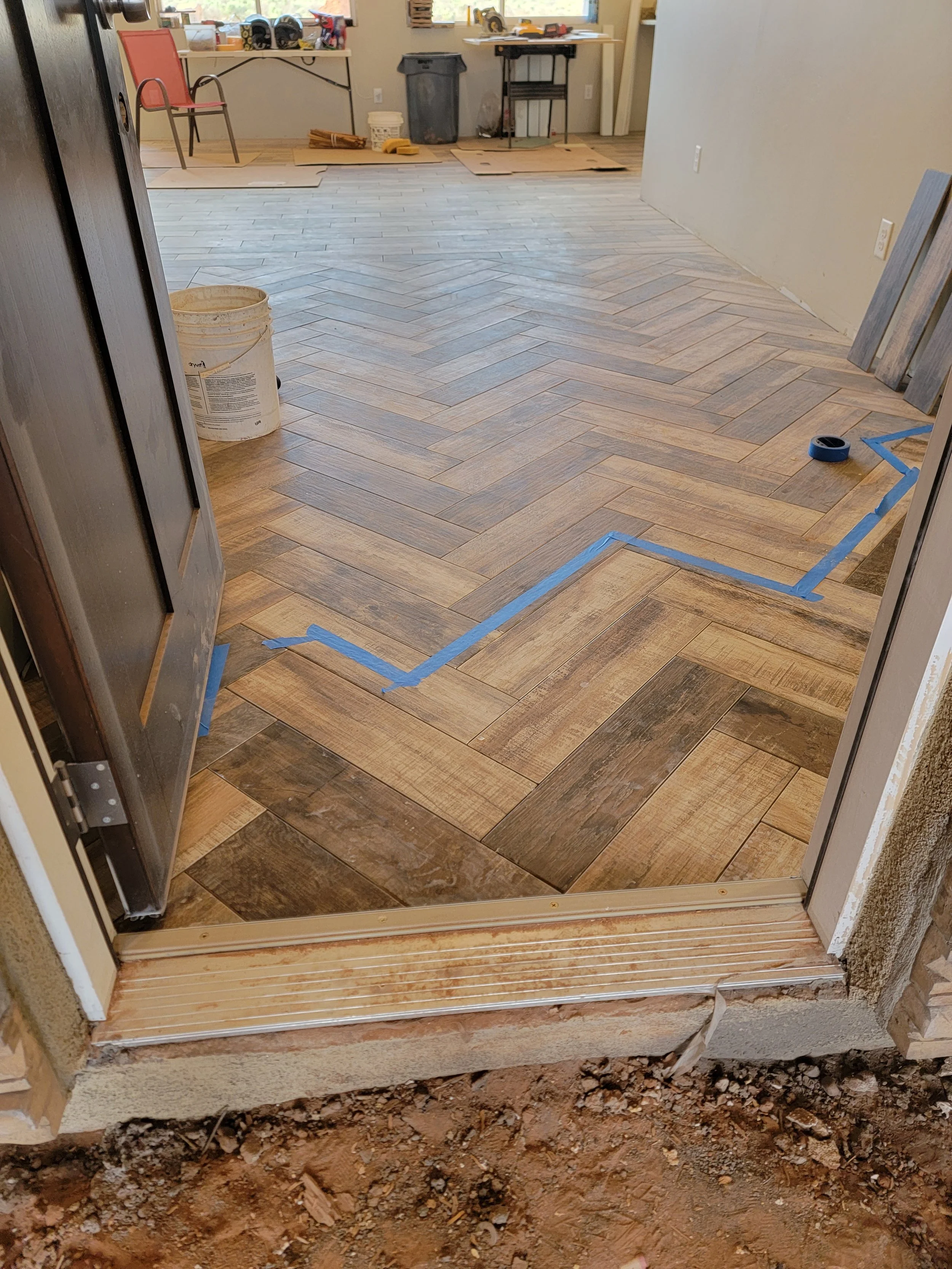 Construction site showing a partially finished wooden flooring in a room, with blue painter's tape outlining the area of new flooring. Tools, construction materials, and a bucket are visible, with an unfinished doorway leading into the room.