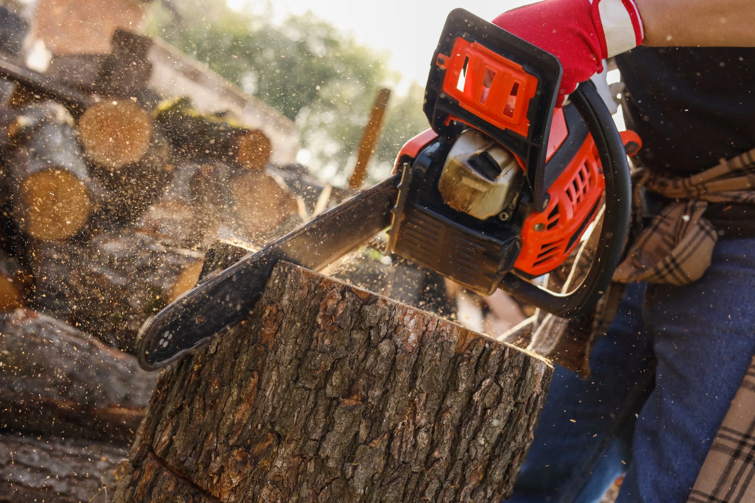 A person wearing red gloves operating an orange chainsaw to cut a log of wood, with wood chips flying in the air.