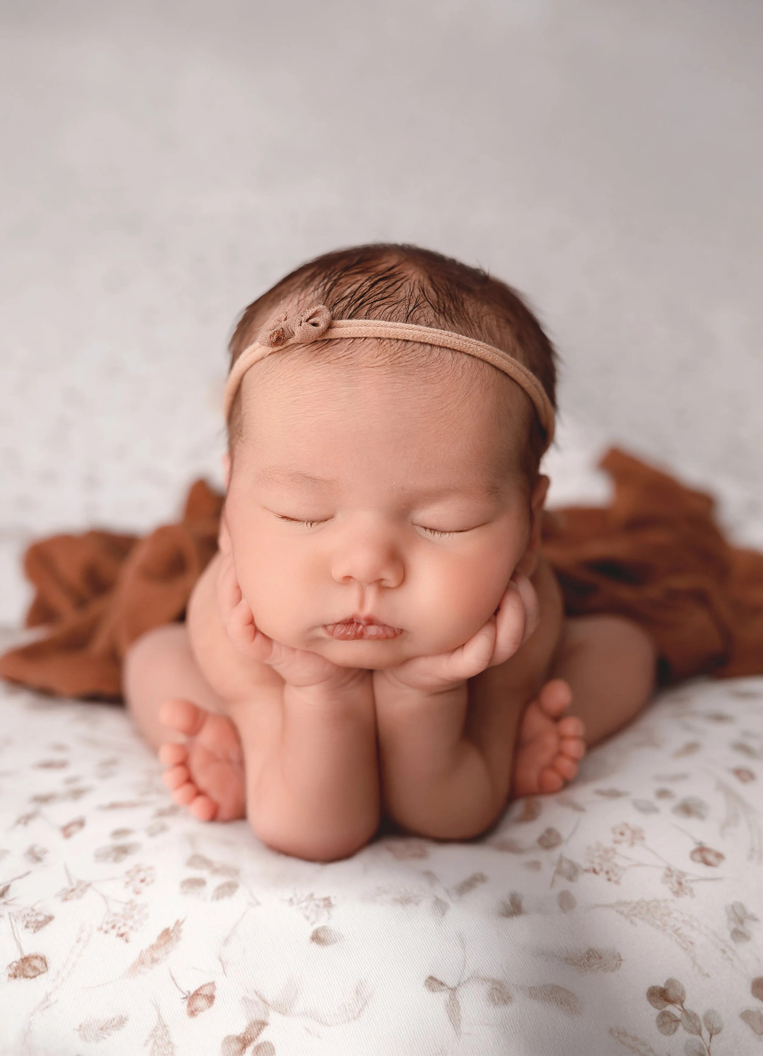 A sleeping baby with a headband, resting on hands with a floral patterned blanket in the background.