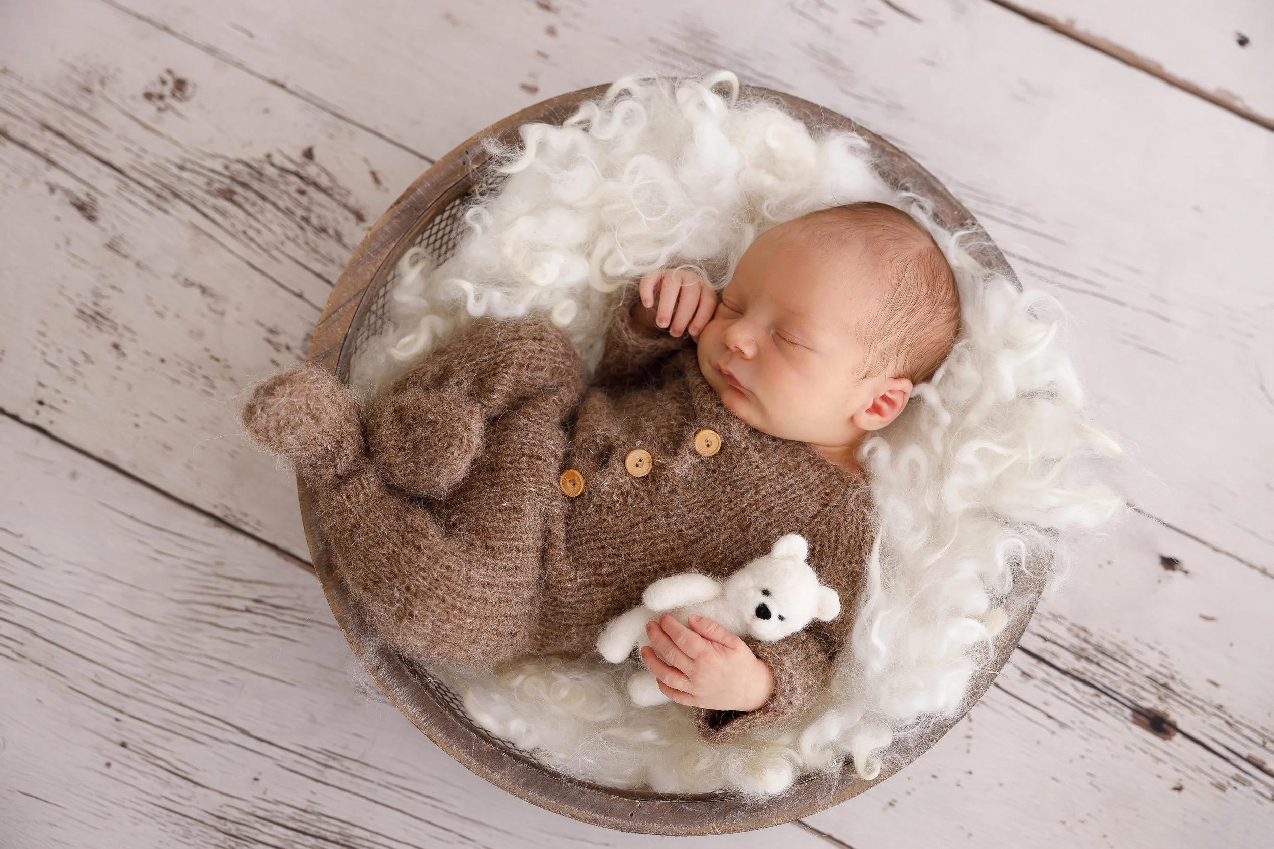 A sleeping baby wrapped in a brown knitted blanket, lying on fluffy white wool inside a round basket on a white wooden floor. The baby has a tiny hand near their face and a small white stuffed bear is near their hand.
