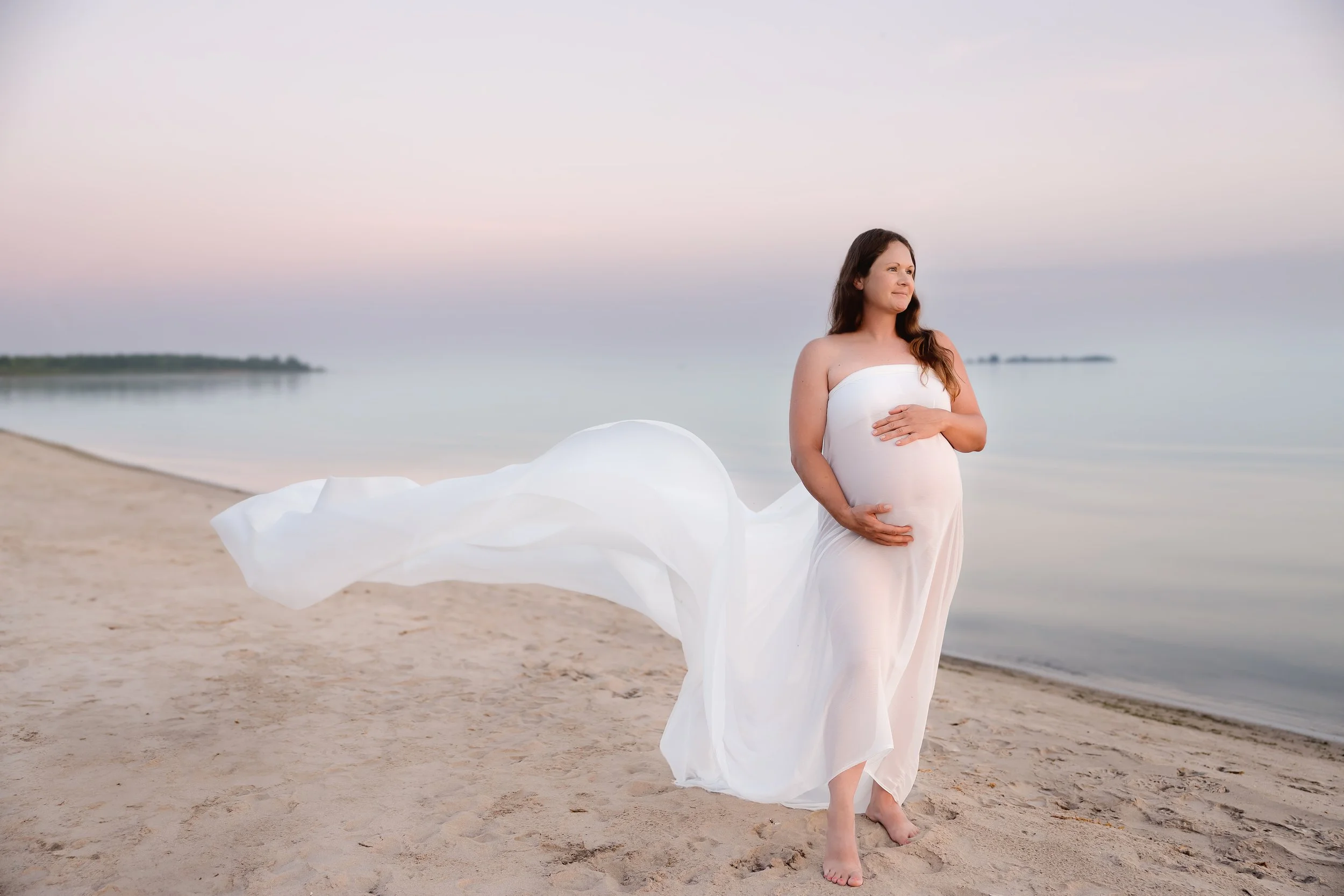 A pregnant woman wearing a white dress, standing barefoot on a sandy beach near calm water during sunset or sunrise, with her hand on her stomach, looking thoughtfully into the distance.