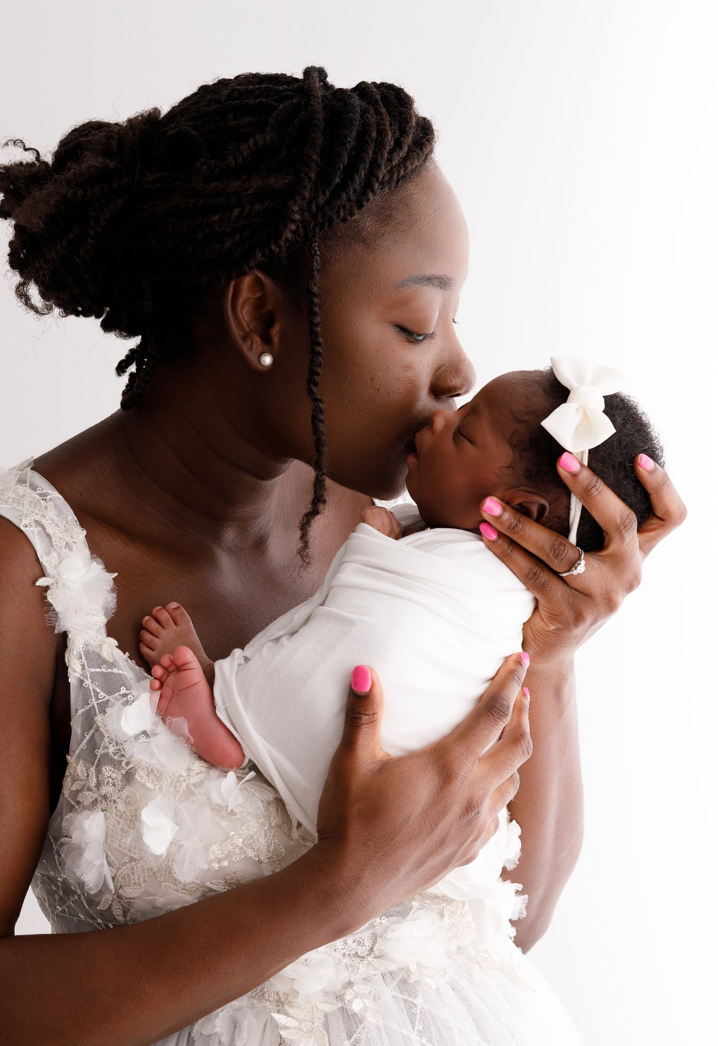 A woman with braided hair, pearl earrings, and pink nail polish is holding a sleeping baby dressed in white with a large white bow in her hair.