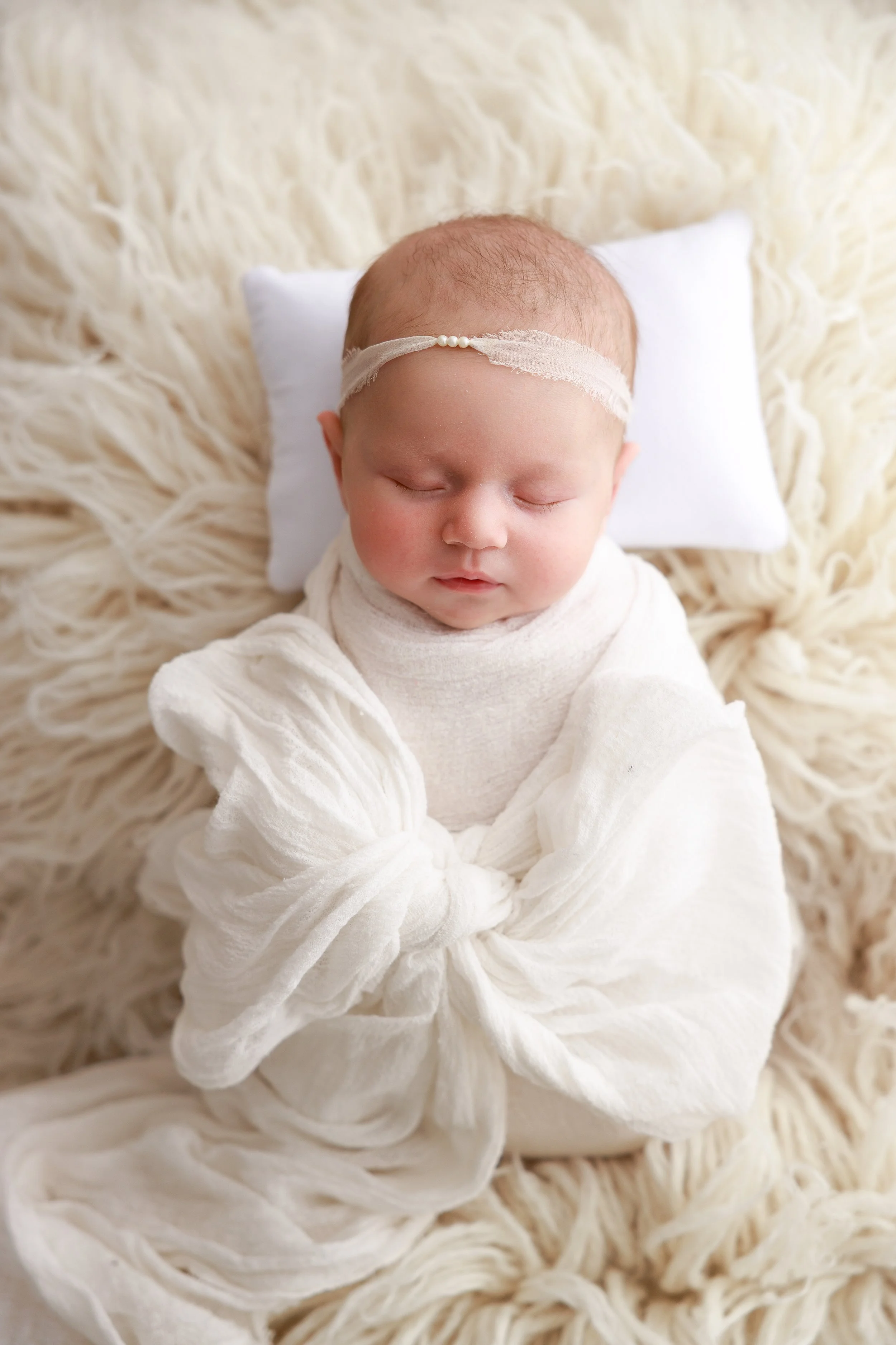 A close-up of a sleeping baby with a lace headband, lying on a white pillow on a fluffy, cream-colored blanket.