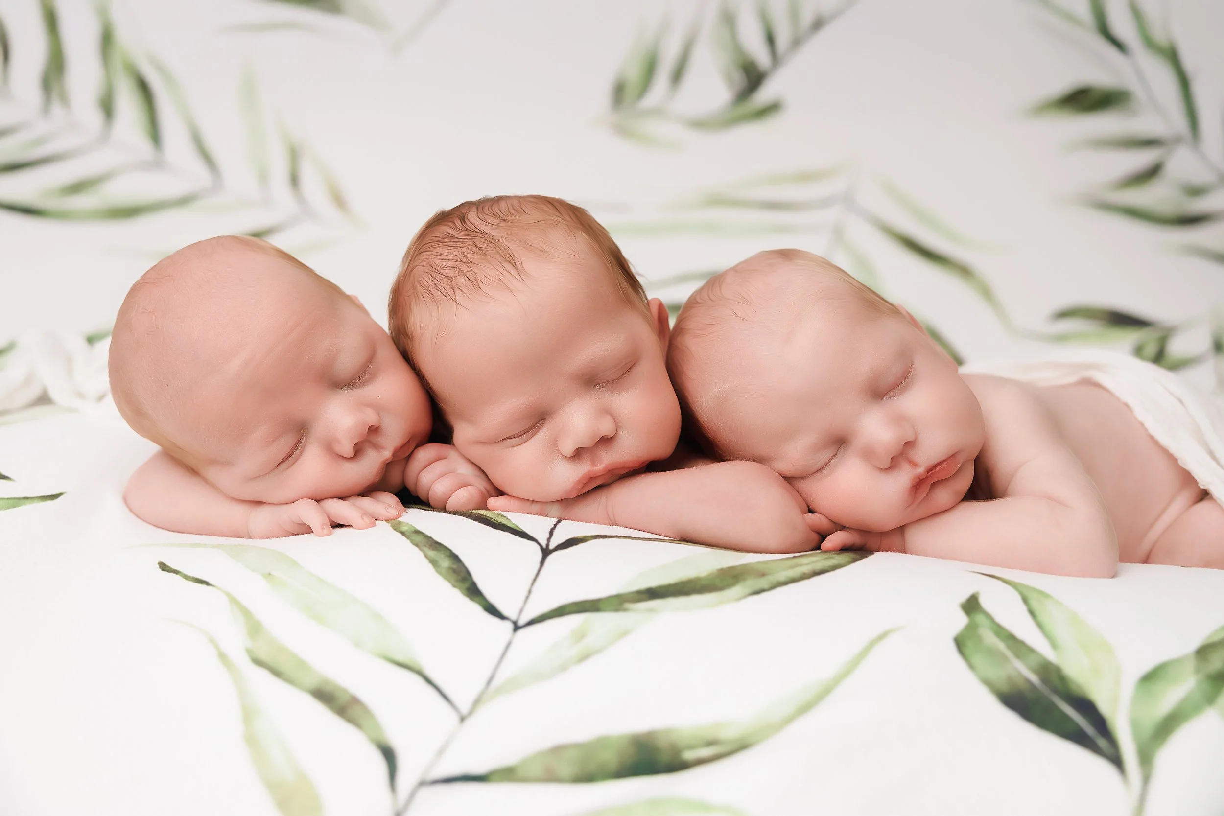 Three sleeping babies lying on a white blanket with green leaf pattern.
