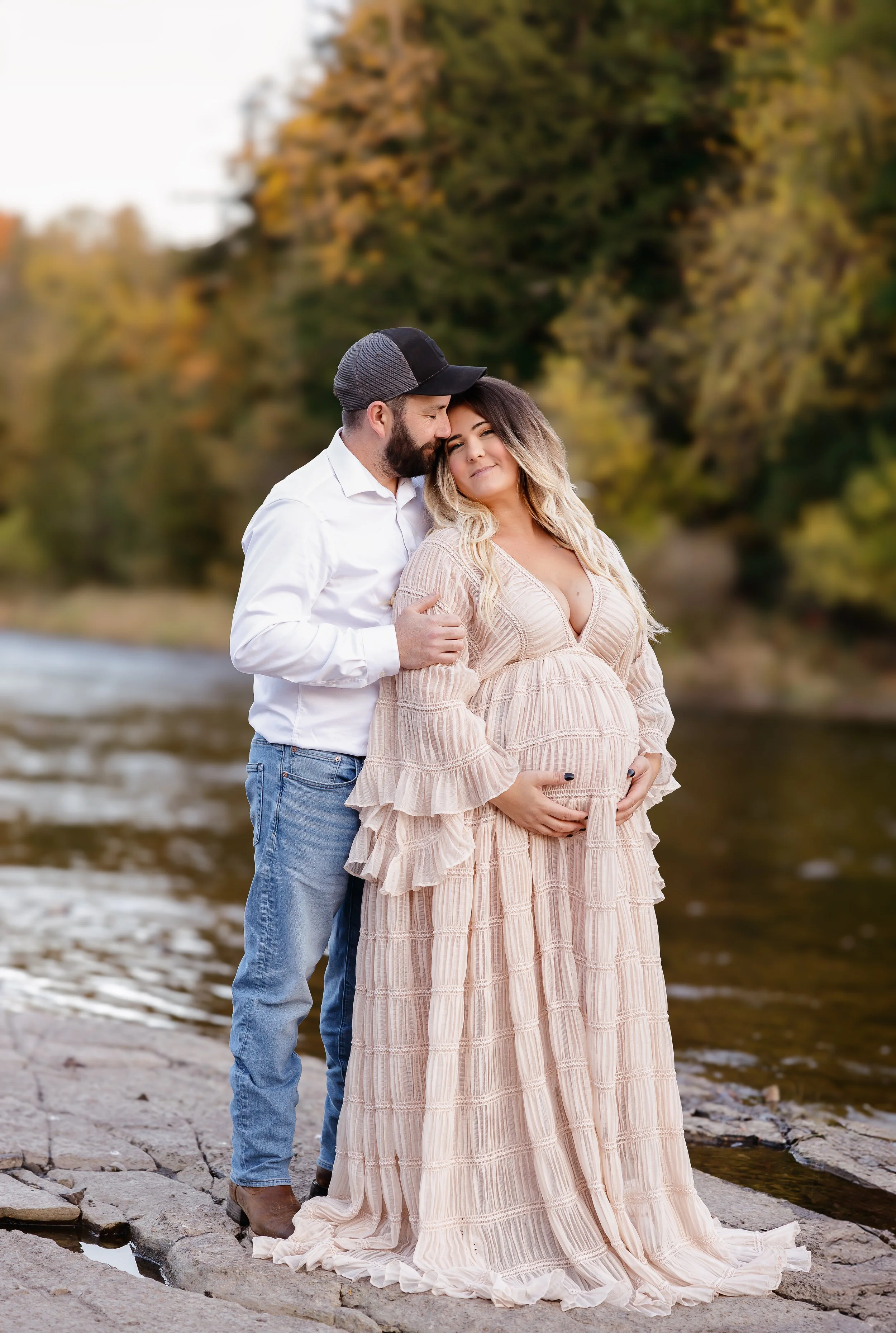 A pregnant woman in a long pink dress standing on a rock by a river, with a man in a white shirt and jeans behind her, gently holding her and kissing her forehead in an outdoor setting with trees in fall colors.