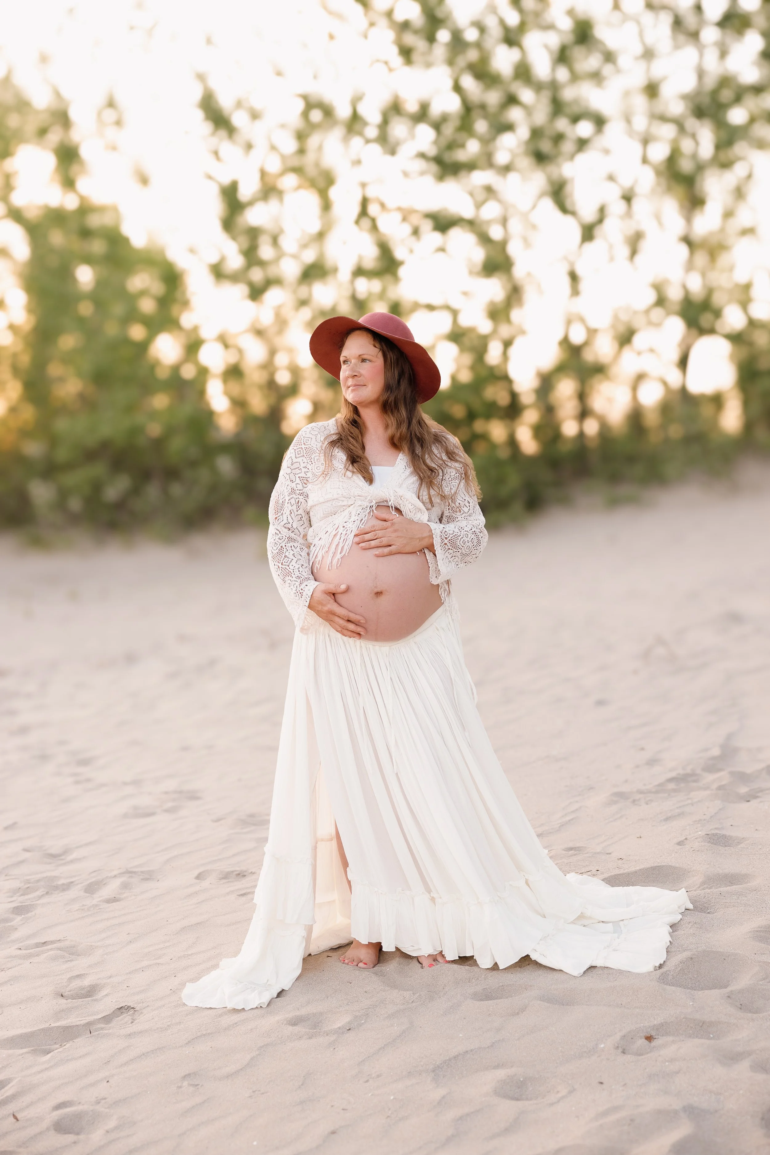 A pregnant woman wearing a white dress and a wide-brimmed hat stands barefoot on sandy ground, holding her belly with one hand and looking into the distance with a serene expression. There are trees with blurred leaves in the background, and the ligh