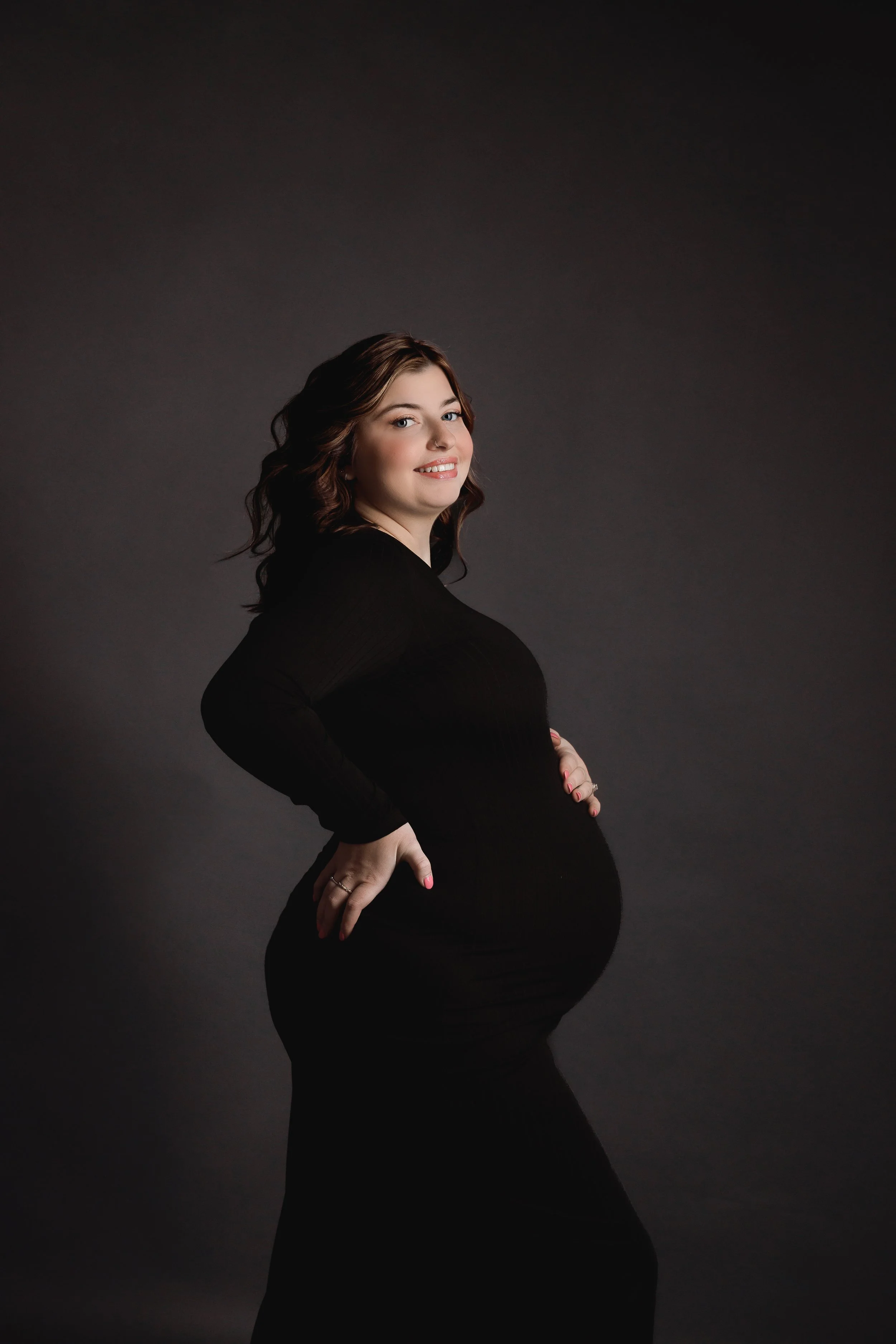 A pregnant woman with brown hair styled in loose waves, smiling and wearing a black long sleeve dress, posing against a dark gray background.