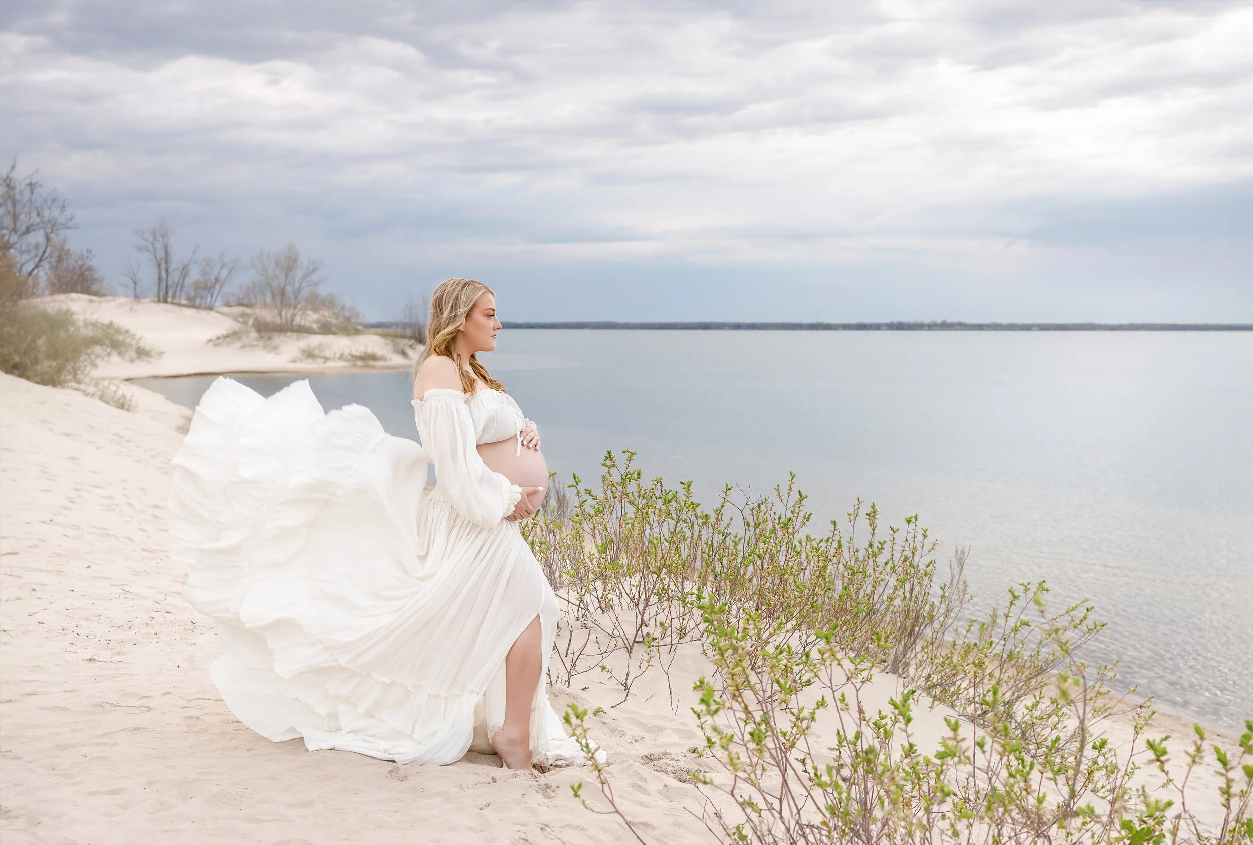 A pregnant woman in a white dress sitting on a sandy beach near water, with a cloudy sky overhead and sparse vegetation around her.