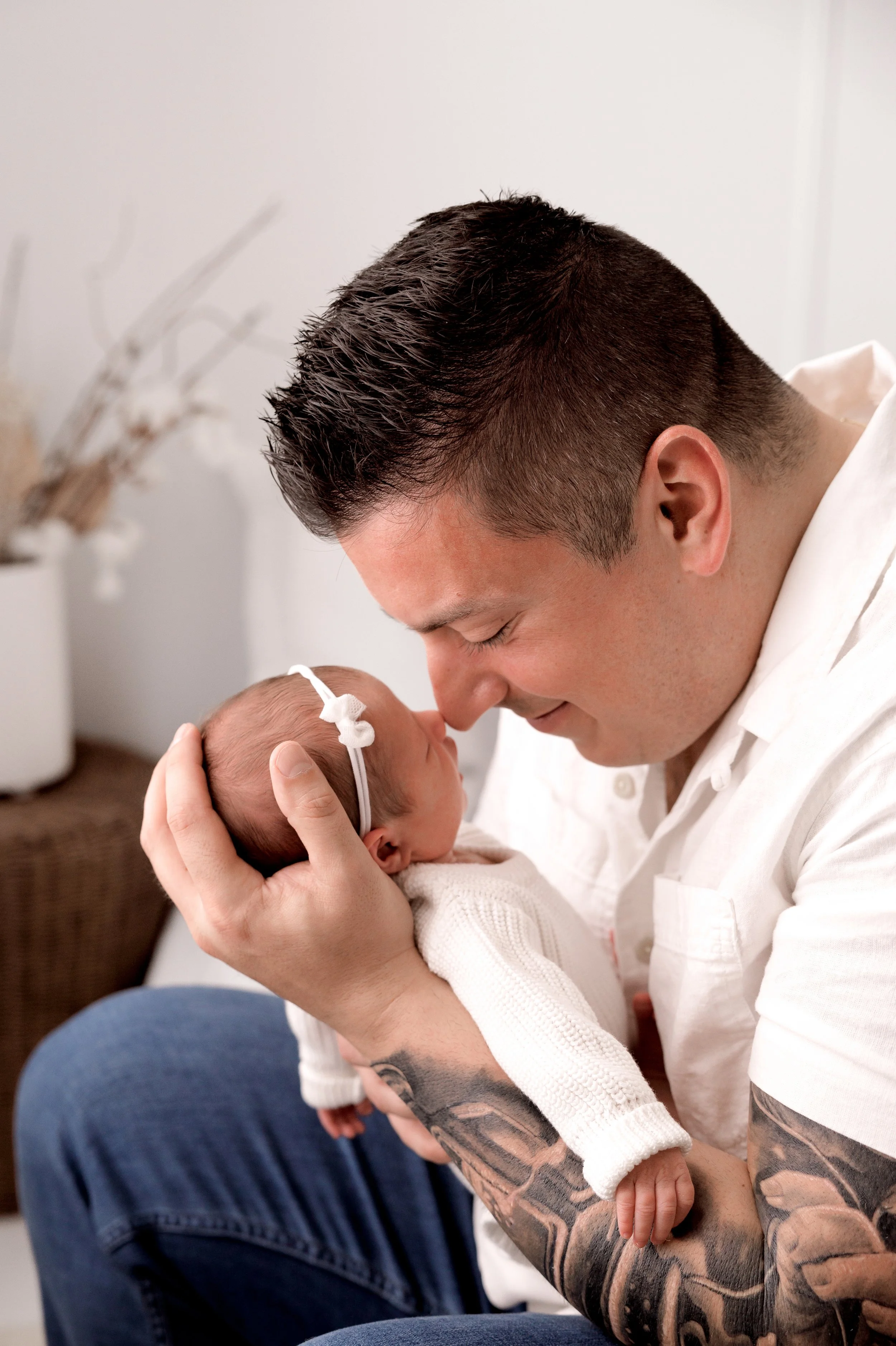 A man with tattoos on his arm gently holds a newborn baby close to his face, both with eyes closed in an intimate moment.