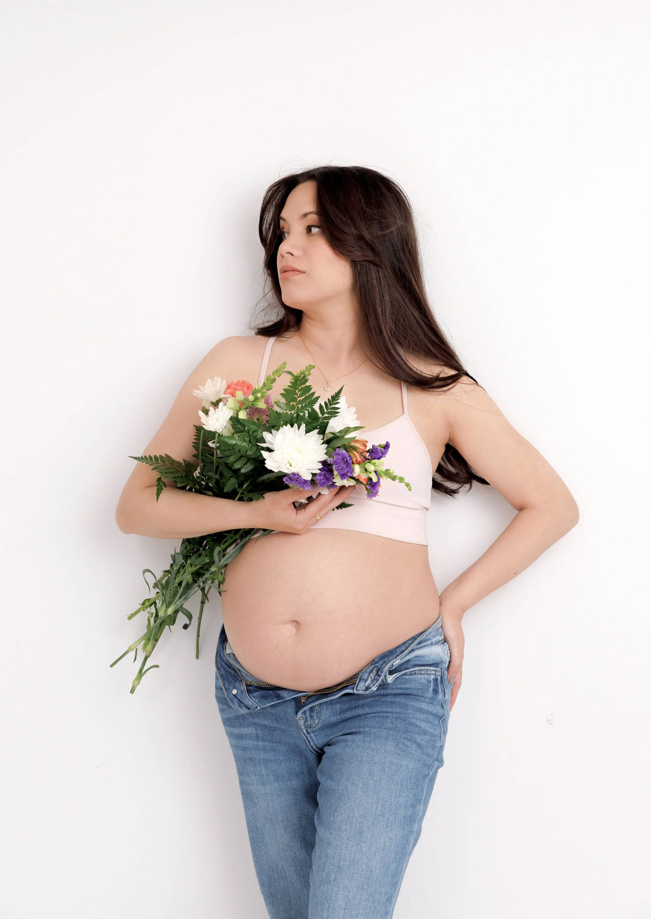 A pregnant woman in casual jeans and a pink bra holding a bouquet of flowers, standing against a white background.